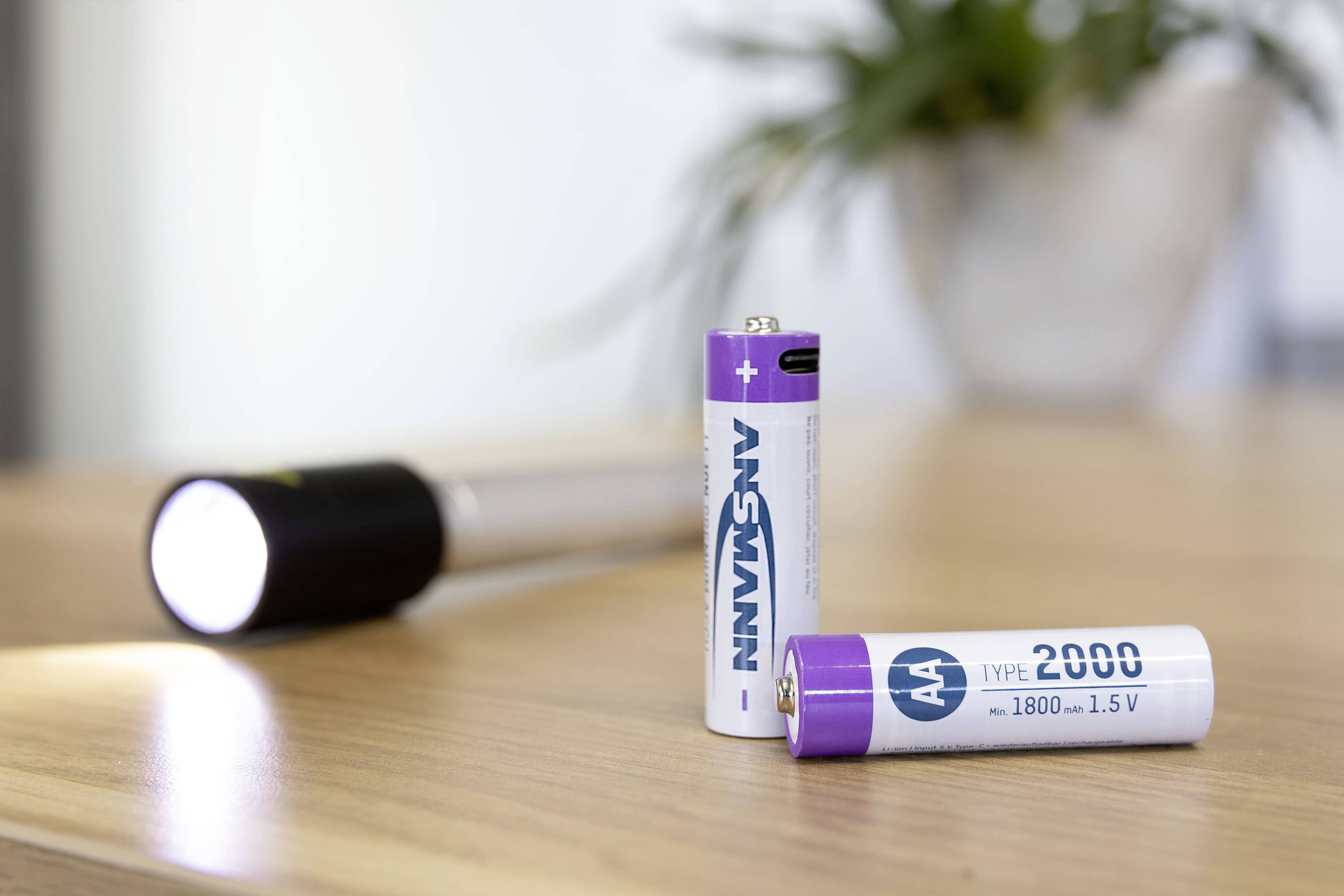 Zwei AA-Batterien stehen auf einem Tisch, eine Taschenlampe im Hintergrund leuchtet. Energierversorgung oder Beleuchtung symbolisiert.