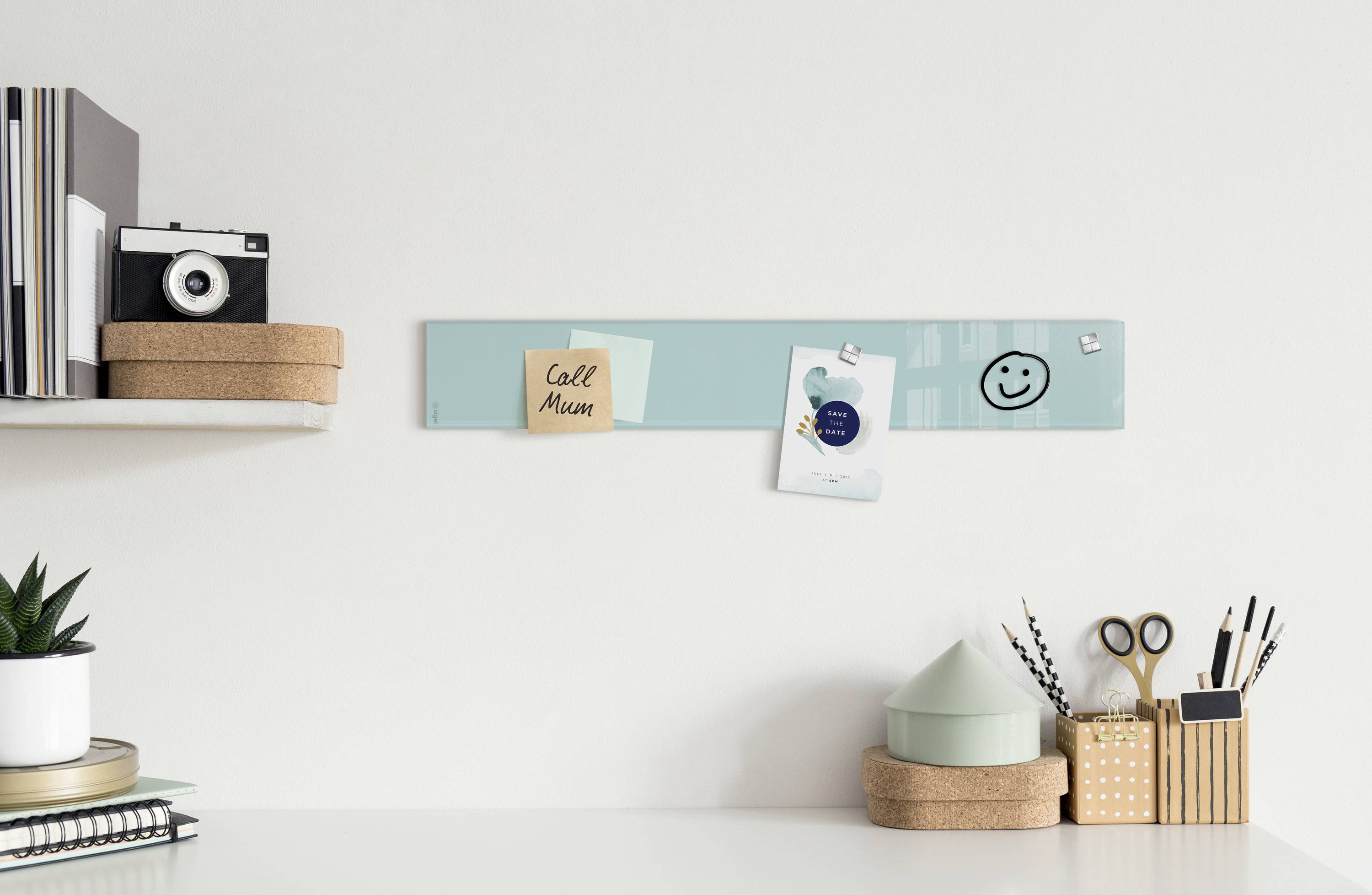 Ein helles Büro mit einem weißen Schreibtisch, auf dem Pflanzen und Schreibwaren stehen. Eine Pinnwand mit Notizen ist an der Wand.