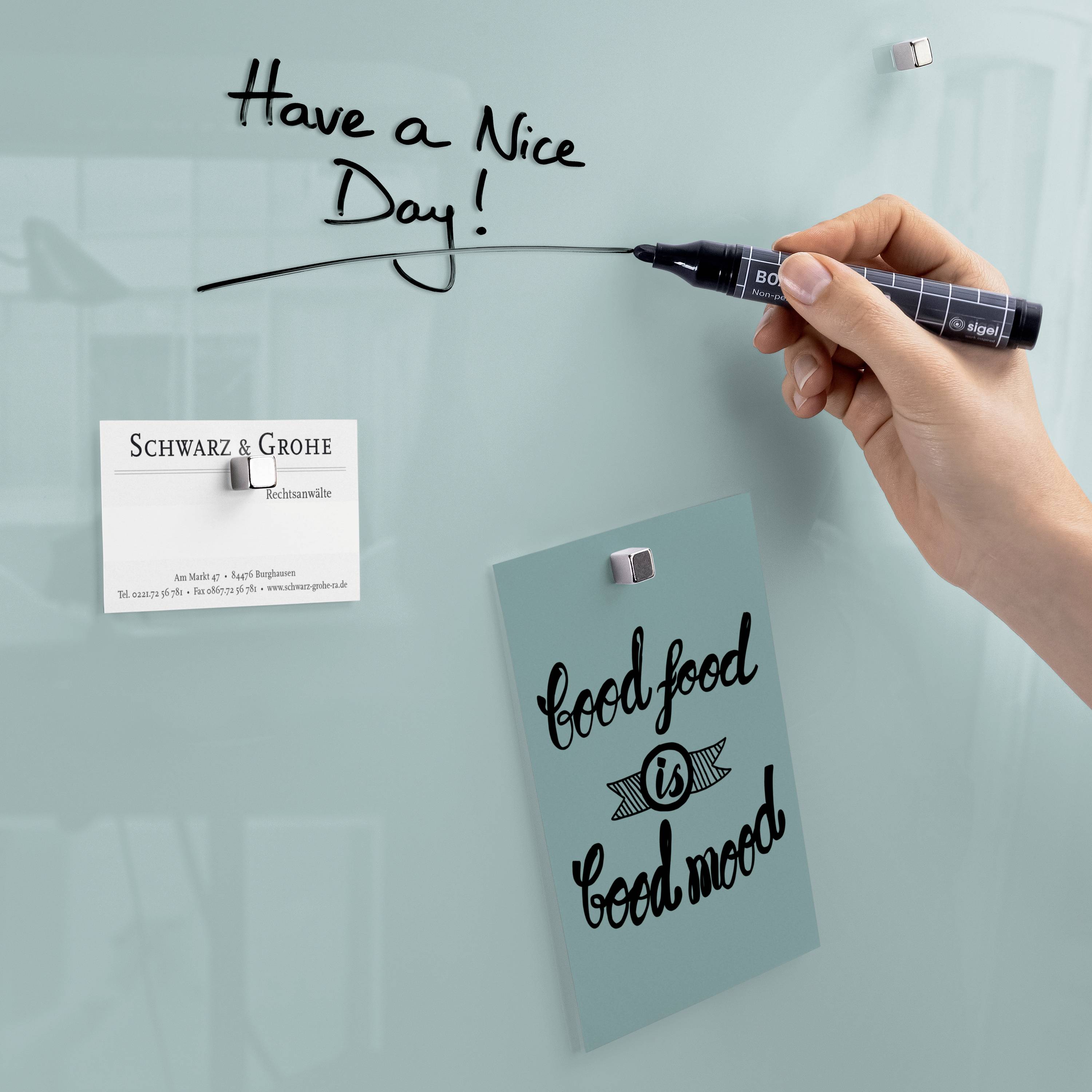 Eine Hand schreibt 'Have a Nice Day' mit einem schwarzen Stift auf eine Glastafel. Daneben hängen ein Zettel mit der Aufschrift 'good food is good mood' und eine Visitenkarte.