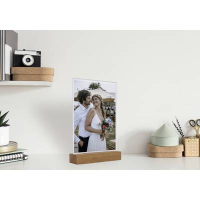 Ein Hochzeitsfoto auf einem Holzrahmen zeigt ein lächelndes Paar, wobei der Mann die Frau umarmt. Im Hintergrund sind Bäume und Zelte sichtbar.
