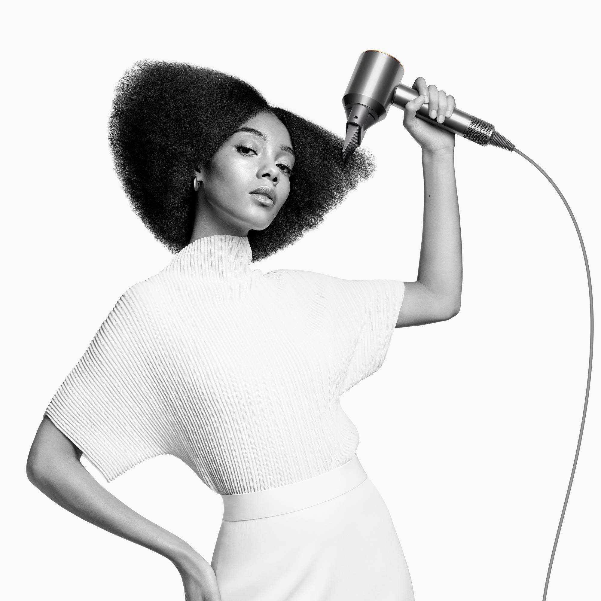 Eine Frau mit stilvollem Afro frischt ihr Haar mit einem Haartrockner auf. Sie trägt ein weißes Oberteil und blickt selbstbewusst.