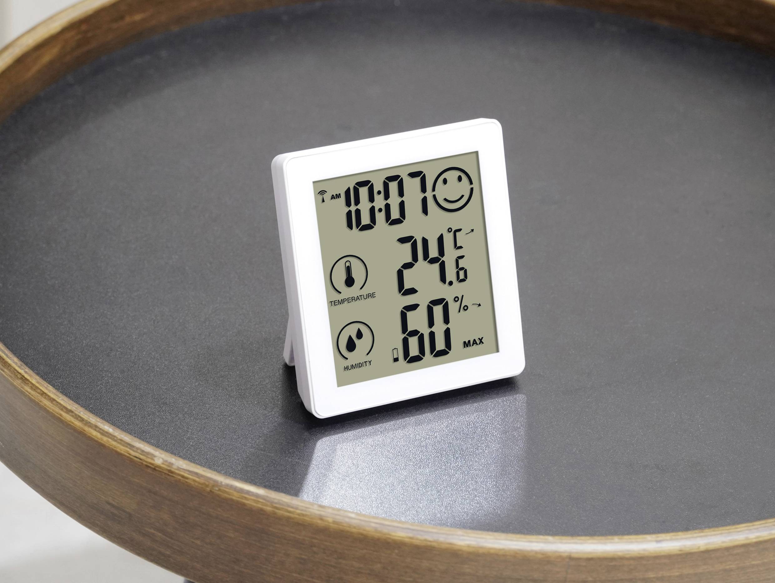 Digitales Thermometer auf Tisch zeigt 24,6°C und 60% Luftfeuchtigkeit an. Lächelndes Gesichtssymbol und aktuelle Uhrzeit: 10:07.