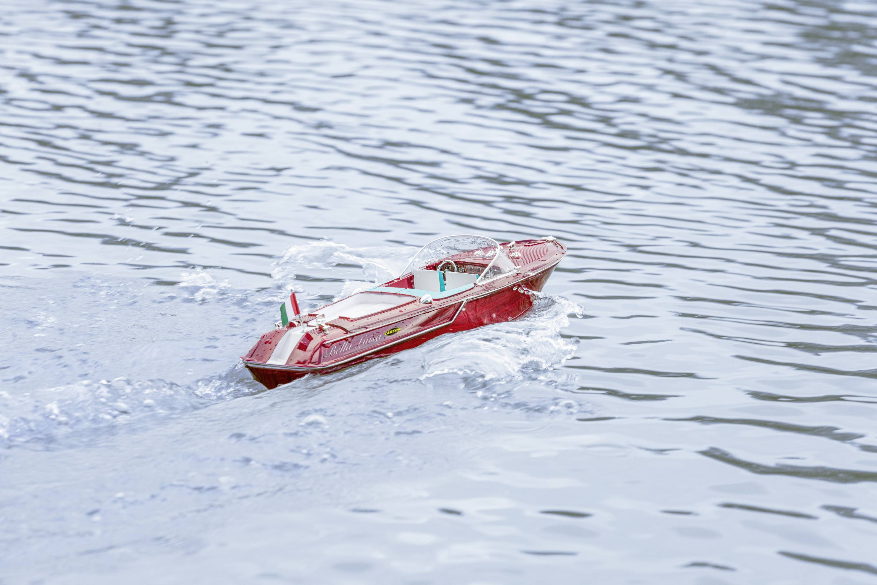 Ein kleines rotes Modellboot fährt auf einem See mit leicht gekräuseltem Wasser. Das Boot erzeugt kleine Wellen während der Fahrt.