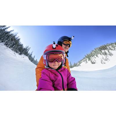 'Zwei Personen in Skiausrüstung fahren bei sonnigem Wetter eine verschneite Piste hinunter, umgeben von schneebedeckten Bäumen.'