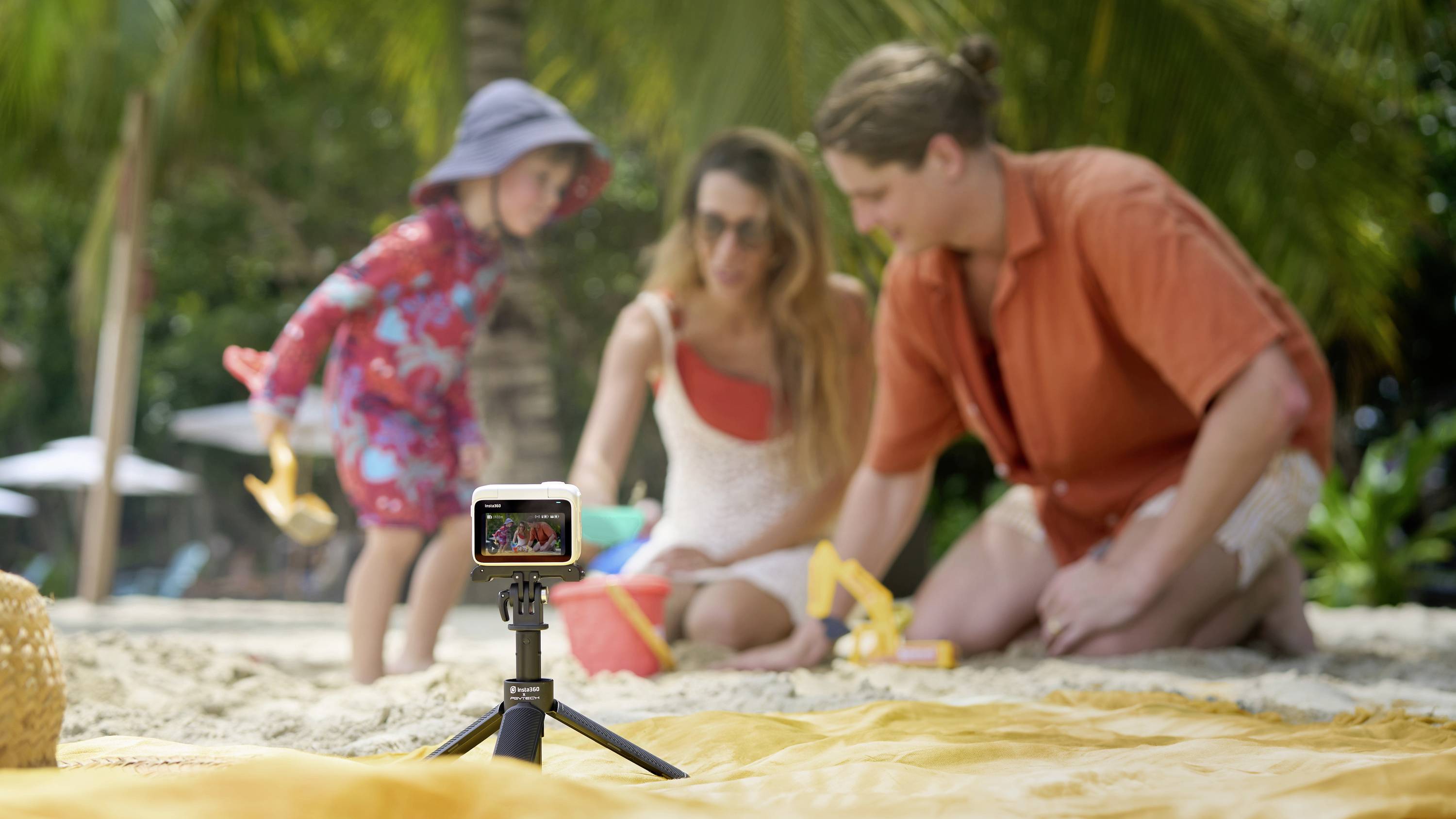 Eine Familie spielt am Strand im Sand. Im Vordergrund steht eine Kamera auf einem Stativ, die die Szene filmt.