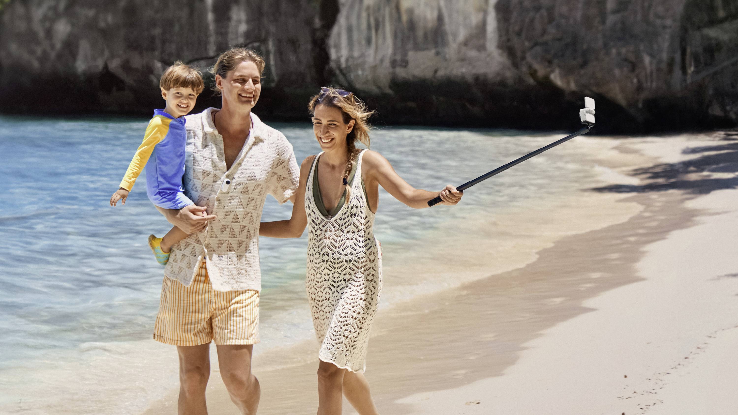 Ein Paar spaziert am Strand, der Mann trägt ein Kind. Die Frau hält einen Selfie-Stick. Im Hintergrund Meer und Felsen.