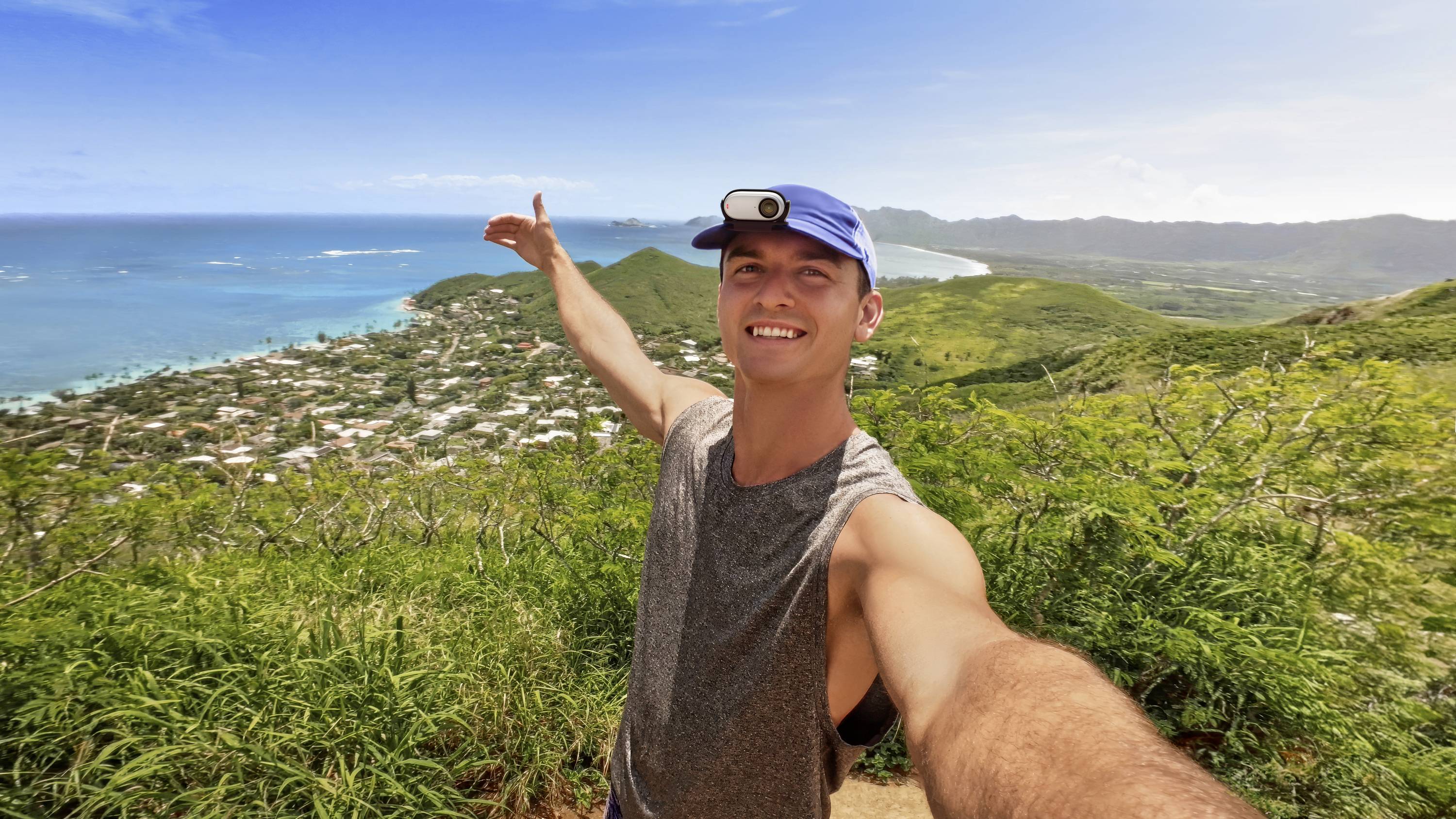 Ein lachender Mann macht ein Selfie auf einem Berg mit Blick auf eine Küstenlandschaft und das Meer im Hintergrund.