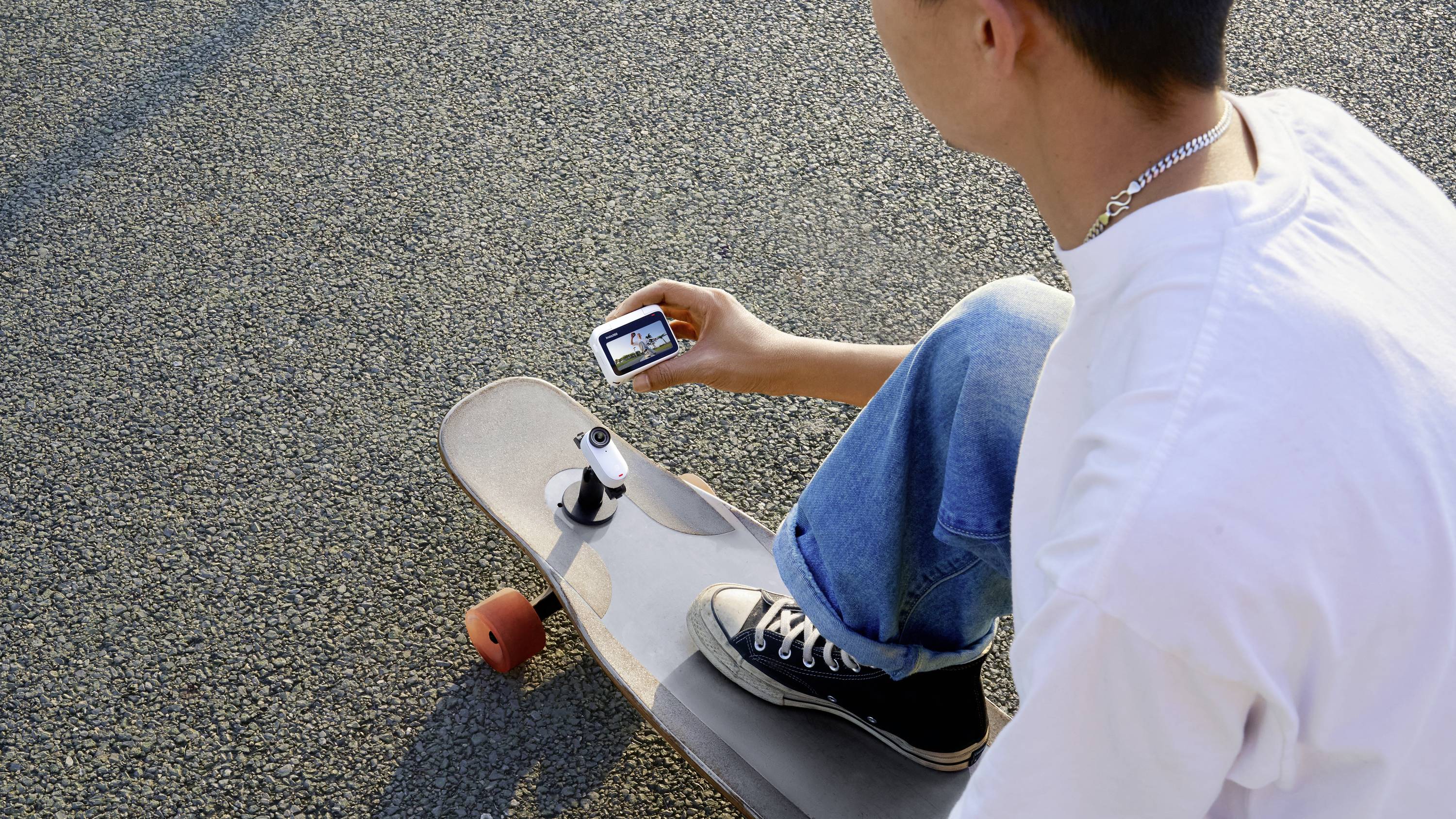 Ein skater in Jeans und weißen T-Shirt prüft ein auf einem Skateboard befestigtes Gerät, vermutlich eine Kamera, auf einem Asphaltweg.