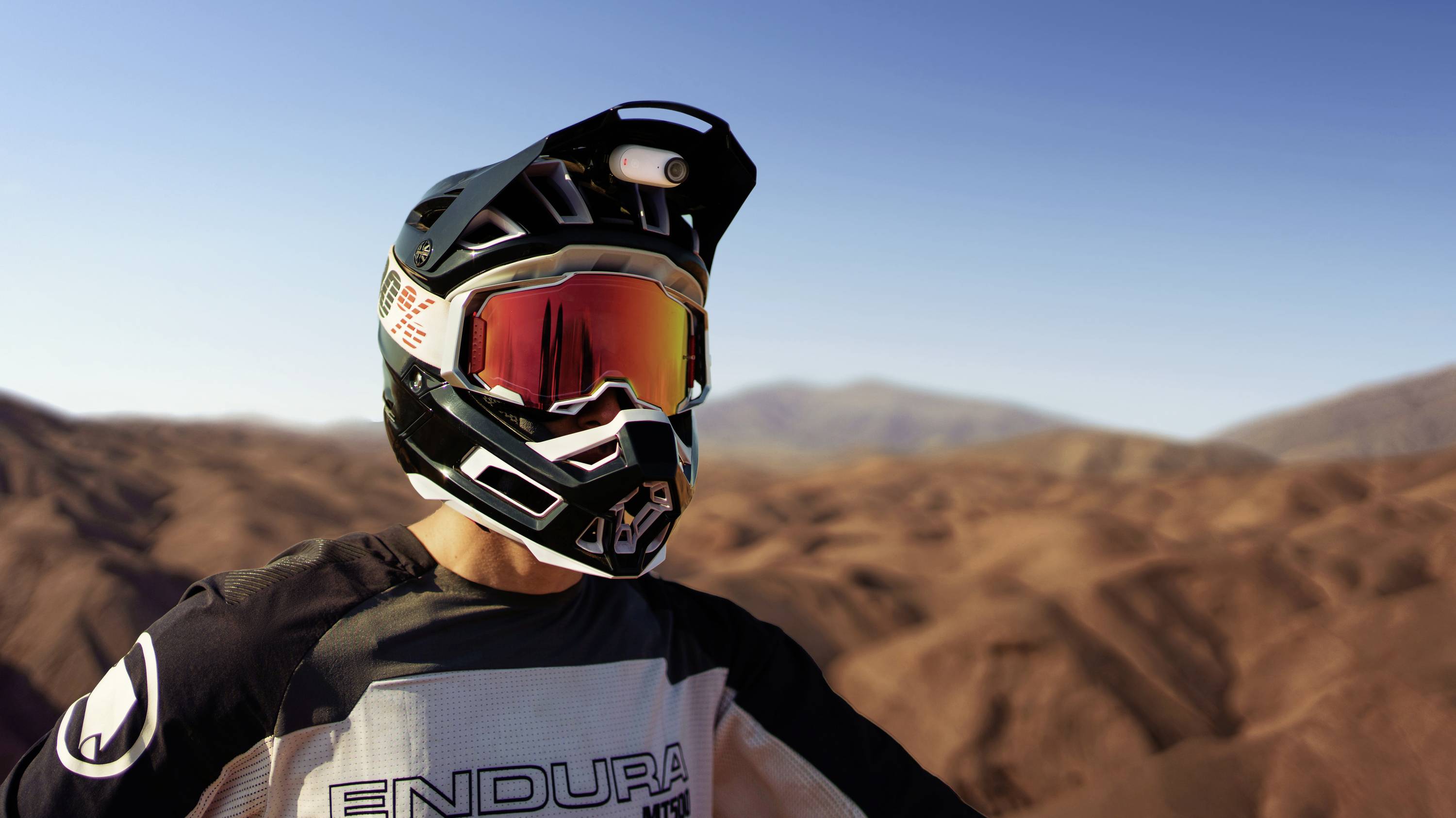Person mit Helm und Brille steht in einer Wüstenlandschaft. Der Helm hat eine Kamera montiert. Im Hintergrund sind sandige Hügel.