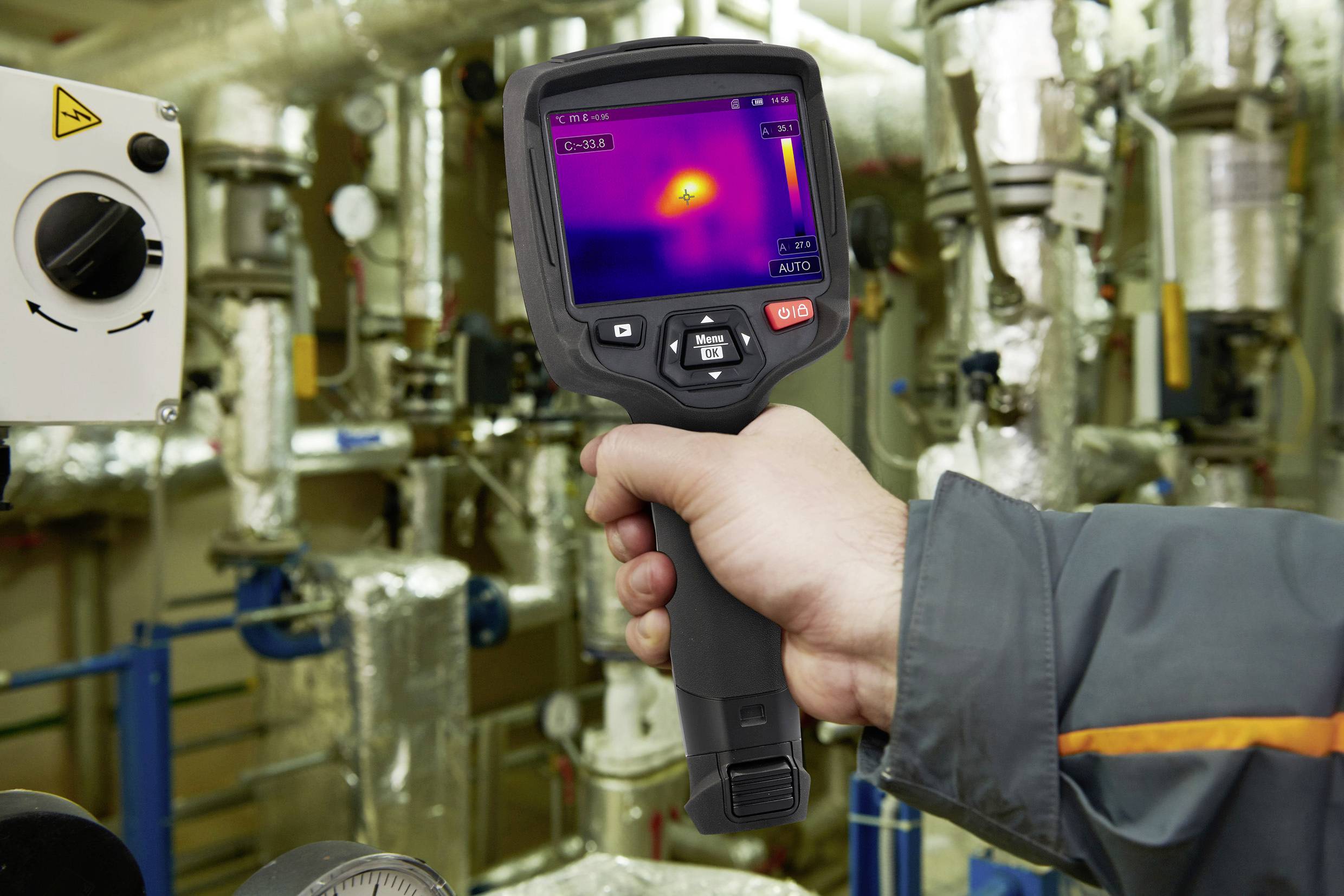 Eine Hand hält eine Wärmebildkamera, die ein Display mit einem Wärmebild anzeigt. Im Hintergrund sind viele Rohre zu sehen.