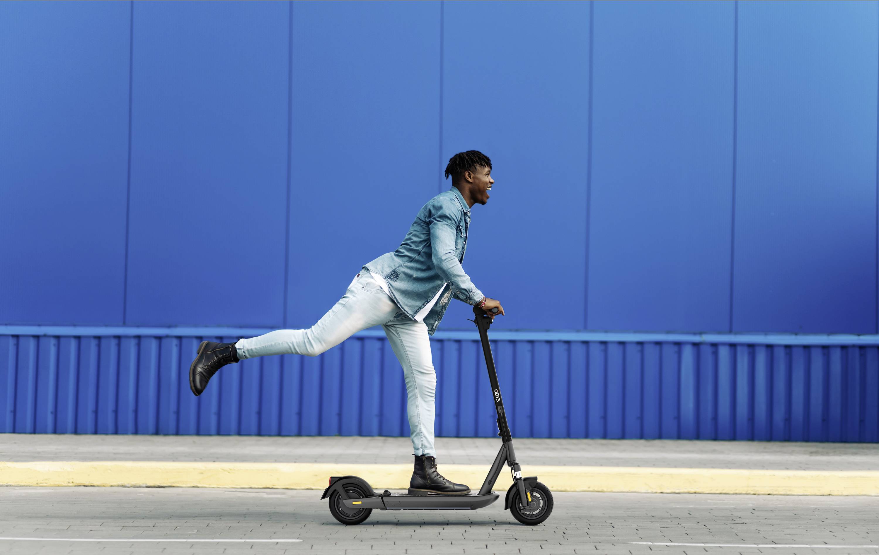 Ein Mann in Jeansjacke und Jeans fährt auf einem Elektroroller vor einer blauen Wand. Sein Bein ist spielerisch in der Luft.