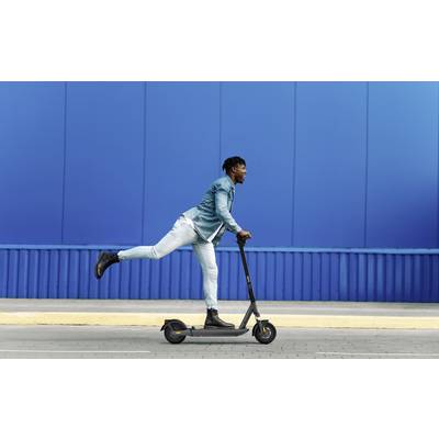 Ein Mann in Jeansjacke und Jeans fährt auf einem Elektroroller vor einer blauen Wand. Sein Bein ist spielerisch in der Luft.