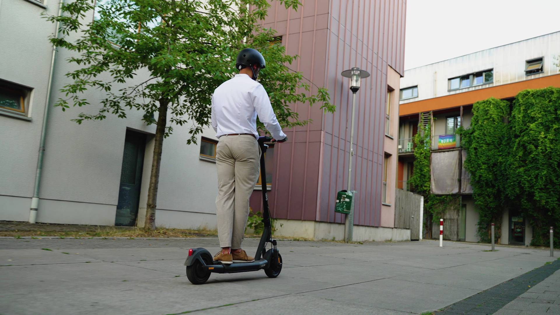 Ein Mann mit Helm fährt auf einem E-Scooter auf einem Bürgersteig in einer Wohngegend. Im Hintergrund sind Gebäude und Bäume zu sehen.