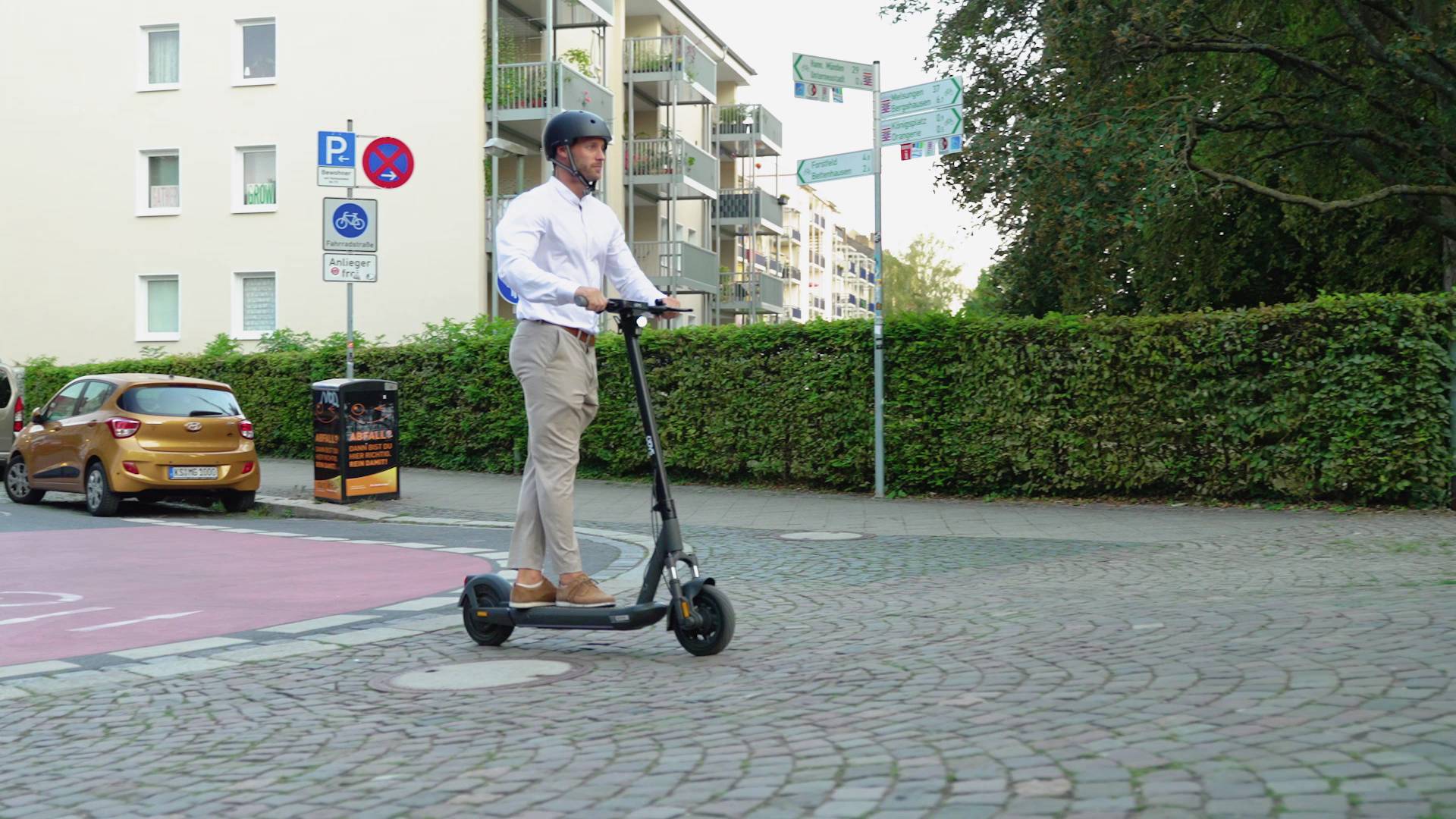 'Person fährt mit einem Elektroroller auf einem Bürgersteig in einer Wohngegend. Wohnungskomplex und Verkehrsschilder im Hintergrund.'