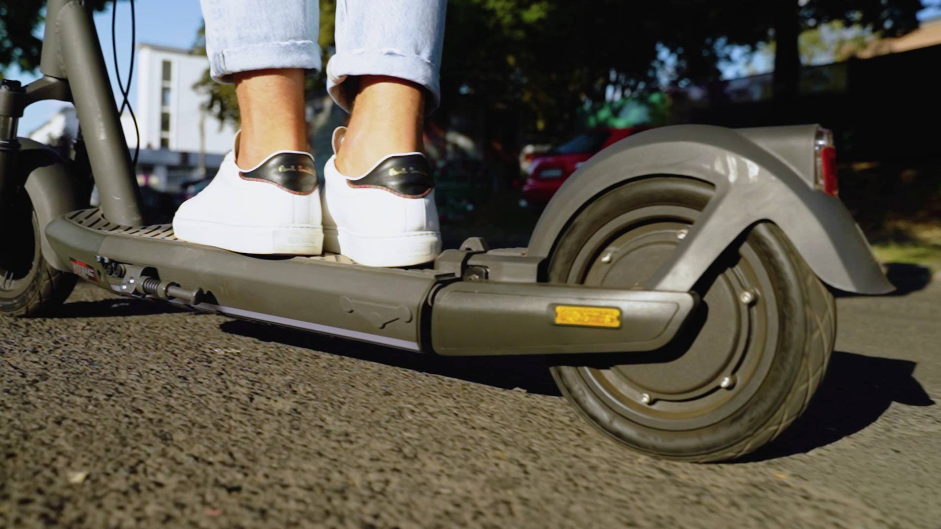 Eine Person steht auf einem Elektroroller und trägt weiße Turnschuhe. Der Roller bewegt sich auf einer asphaltierten Straße.