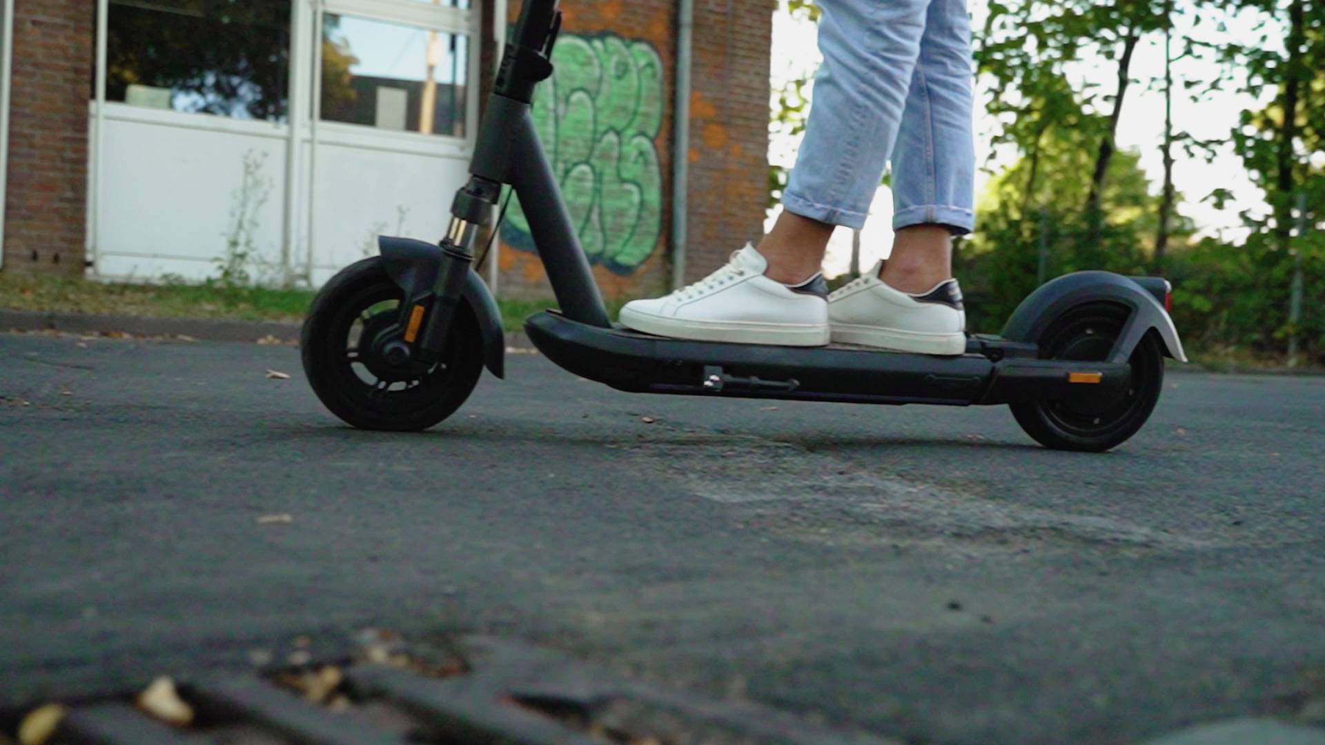 Eine Person fährt einen E-Scooter auf einer leeren Straße. Im Hintergrund sind Bäume und eine Backsteinmauer zu sehen.