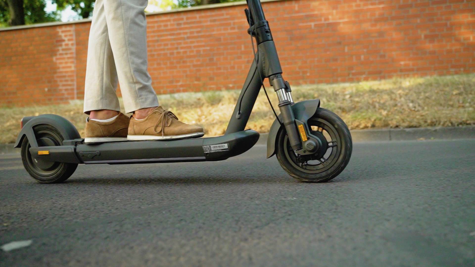 Person fährt mit einem E-Scooter auf einer Straße vor einer rotbraunen Mauer. Der Fokus liegt auf den Beinen und dem Scooter.