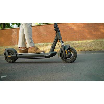 Person fährt mit einem E-Scooter auf einer Straße vor einer rotbraunen Mauer. Der Fokus liegt auf den Beinen und dem Scooter.