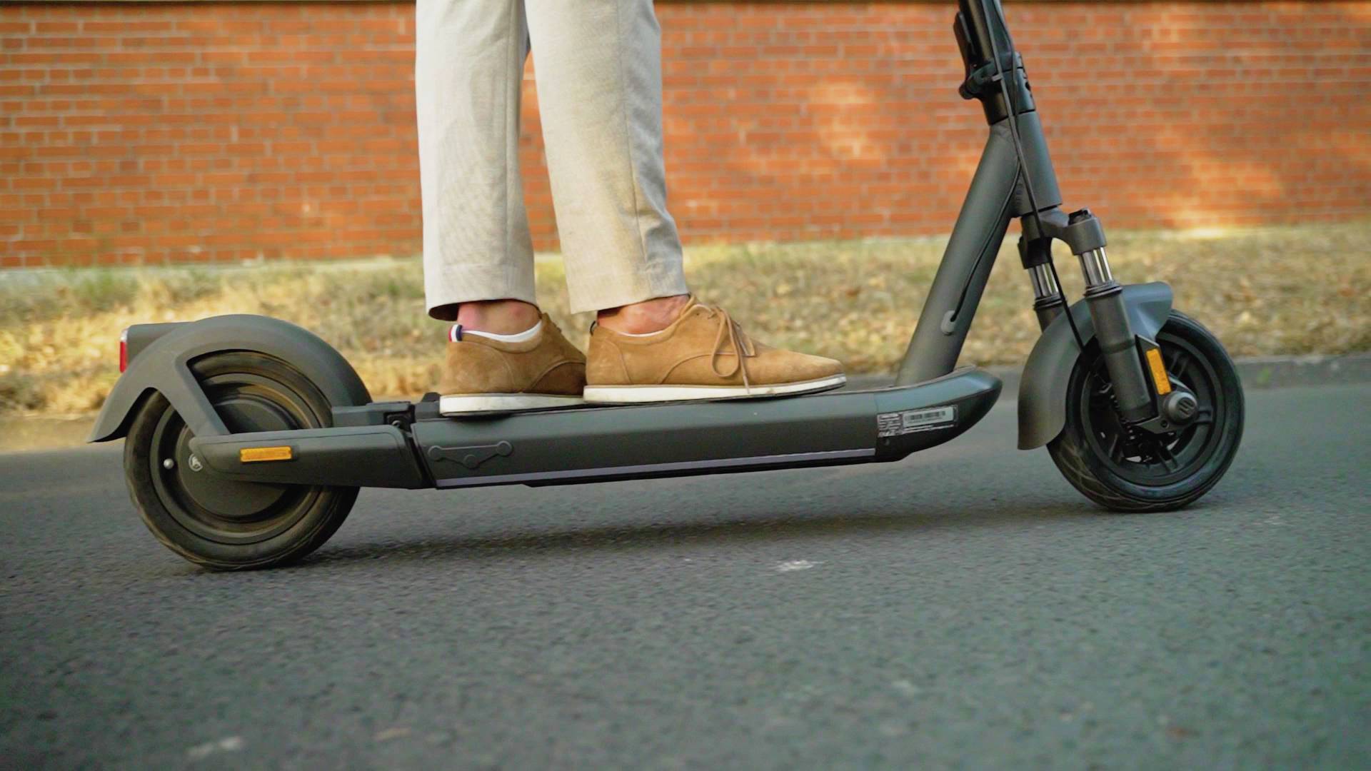 Person auf Elektroroller fährt auf Asphaltstraße, rote Ziegelmauer im Hintergrund.