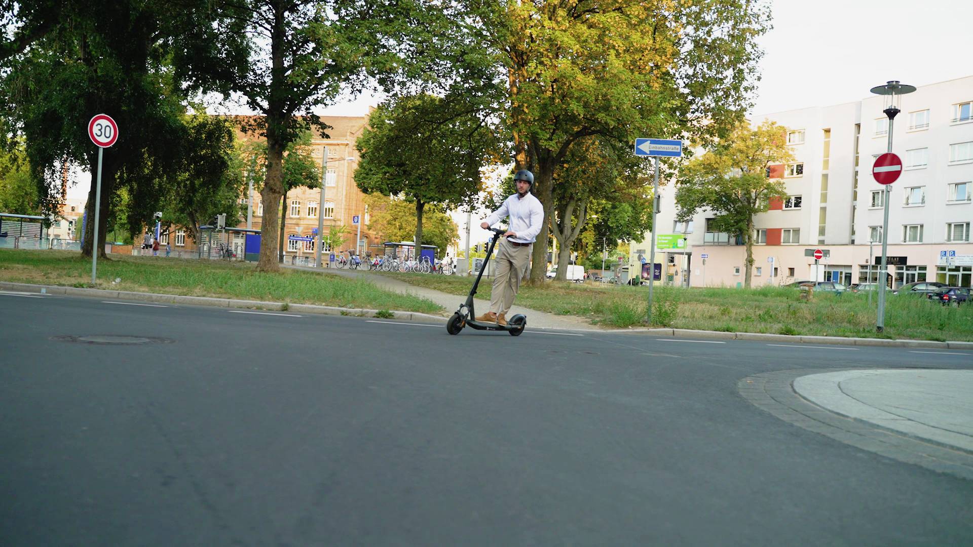 Eine Person fährt bei Tageslicht mit einem E-Scooter auf einer ruhigen Straße vorbei an Wohnhäusern und Verkehrsschildern.