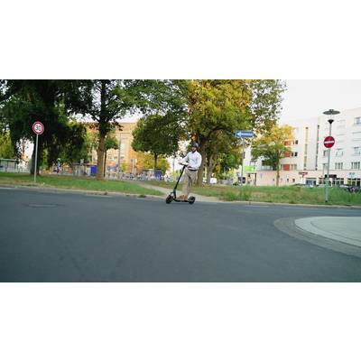 Eine Person fährt bei Tageslicht mit einem E-Scooter auf einer ruhigen Straße vorbei an Wohnhäusern und Verkehrsschildern.