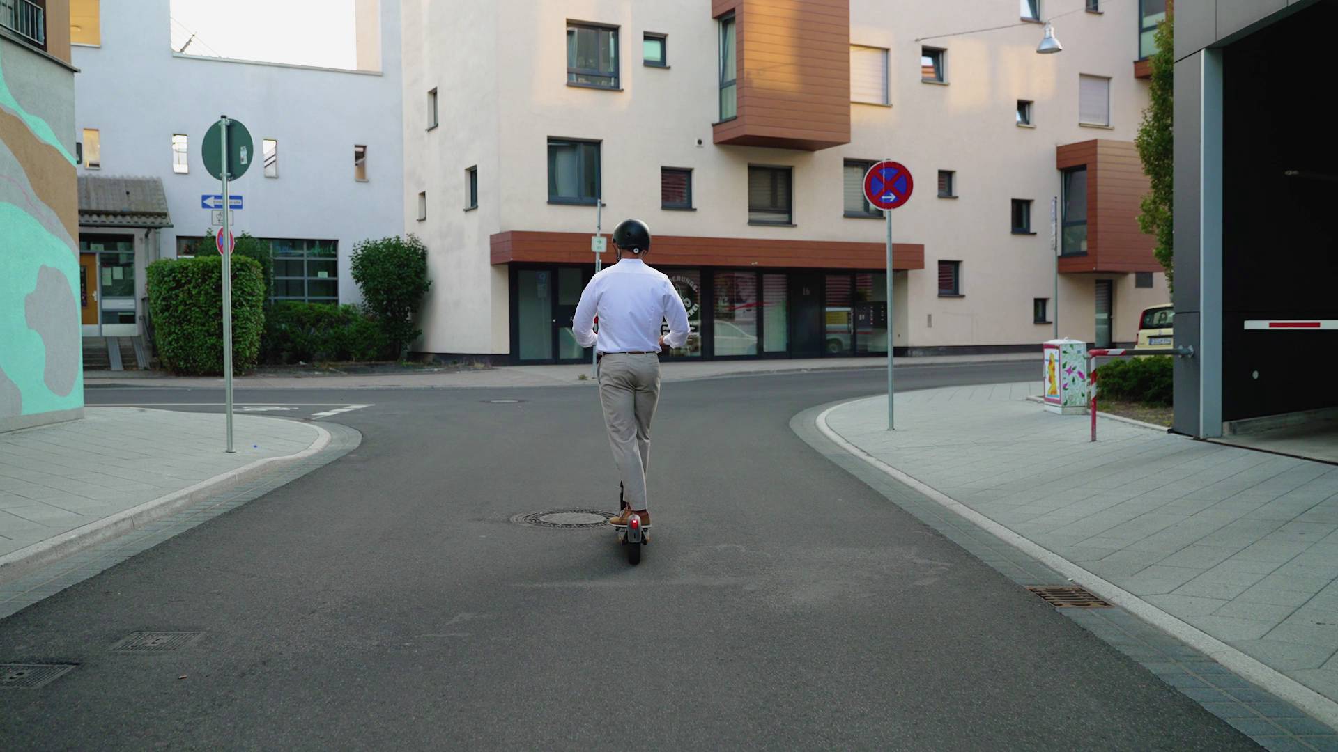 Eine Person fährt einen Elektroroller in einer städtischen Umgebung. Im Hintergrund sind moderne Wohngebäude zu sehen.