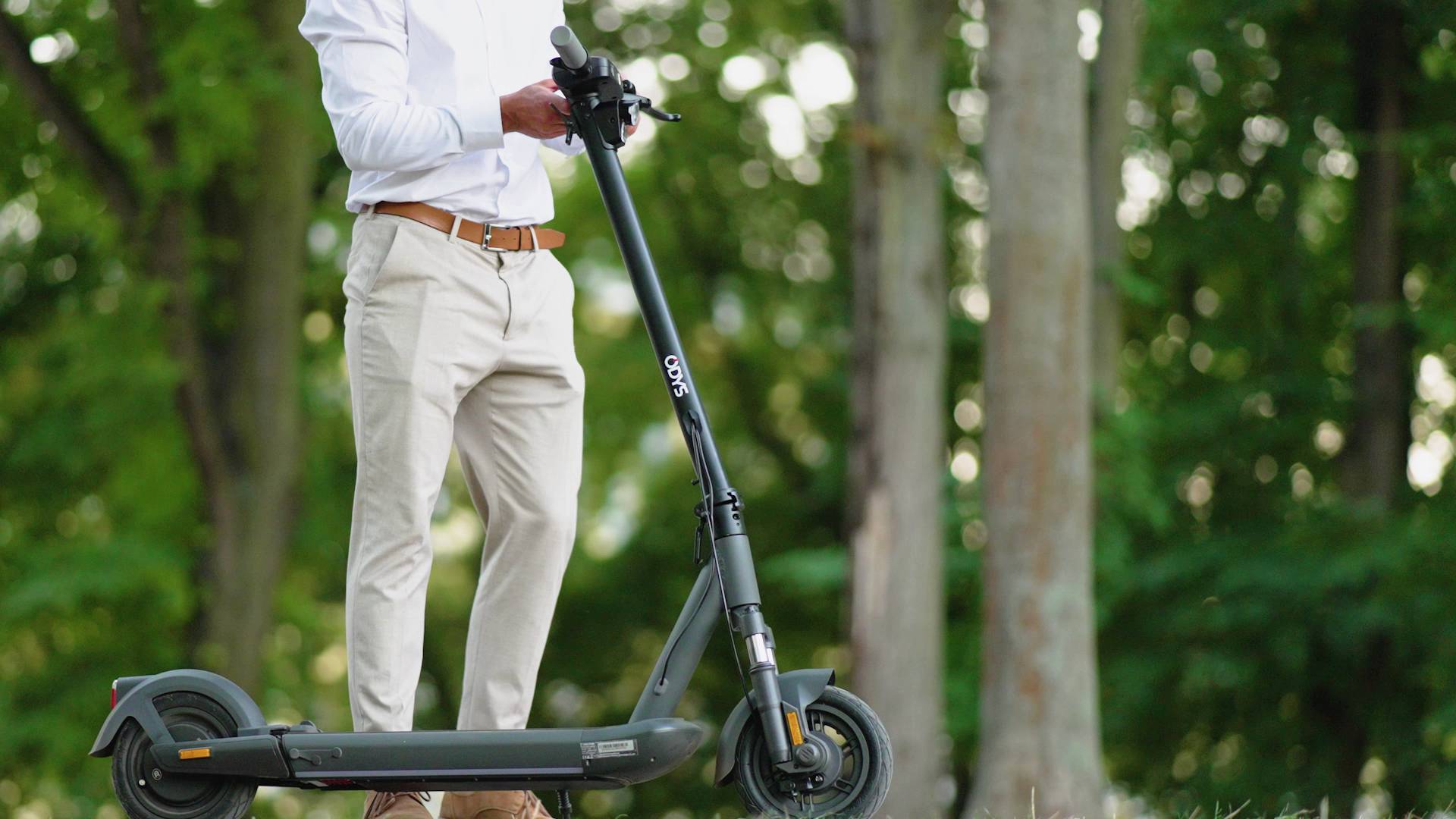 Eine Person in eleganter Kleidung steht auf einem schwarzen E-Scooter in einem grünen, bewaldeten Bereich.