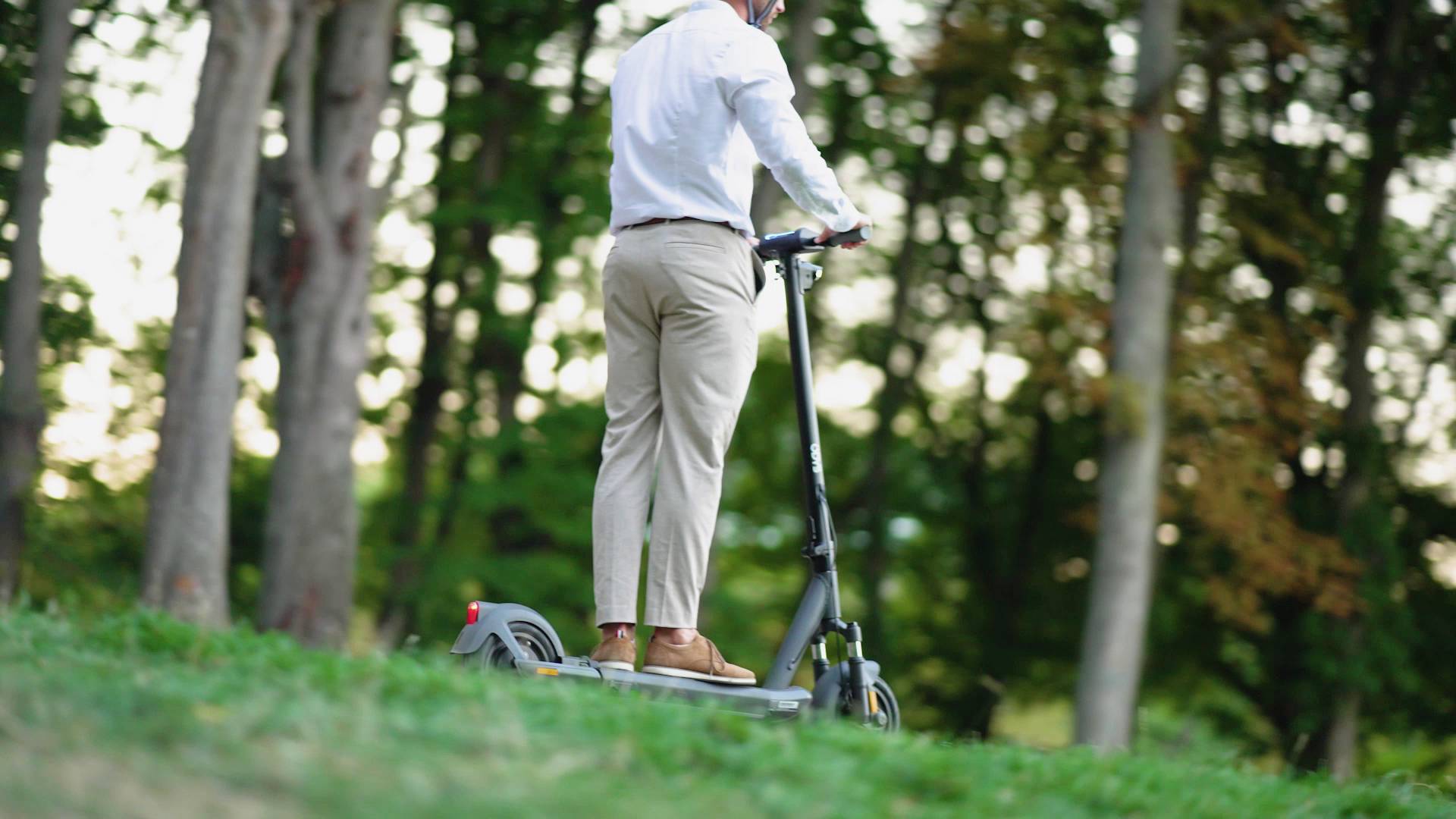 Eine Person fährt einen Elektroroller auf einem grasbedeckten Weg durch einen Wald. Sie trägt ein weißes Hemd und beige Hosen.