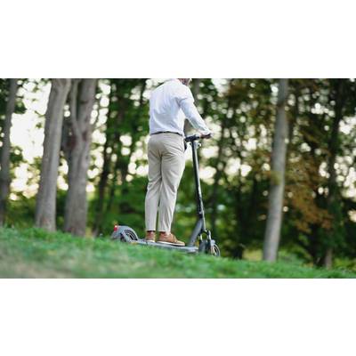 Eine Person fährt einen Elektroroller auf einem grasbedeckten Weg durch einen Wald. Sie trägt ein weißes Hemd und beige Hosen.