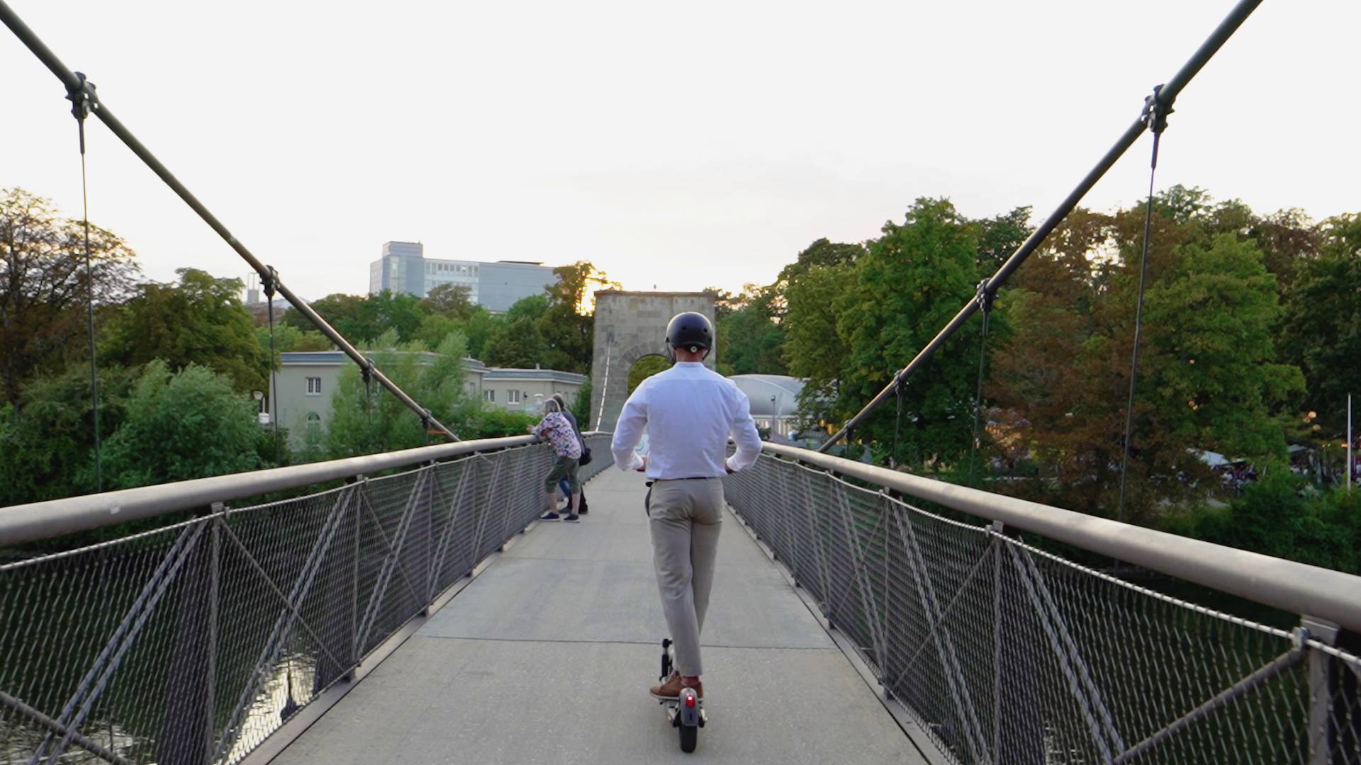Eine Person fährt auf einem Elektro-Scooter über eine Fußgängerbrücke. Im Hintergrund sind Bäume und Gebäude zu sehen.