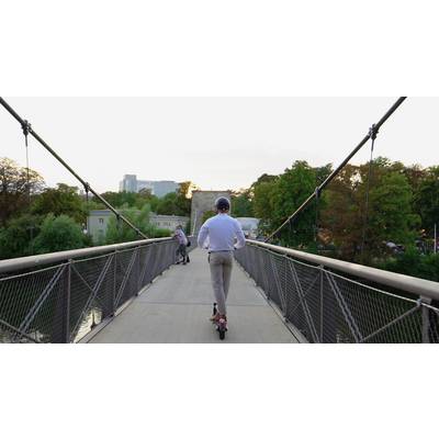 Eine Person fährt auf einem Elektro-Scooter über eine Fußgängerbrücke. Im Hintergrund sind Bäume und Gebäude zu sehen.