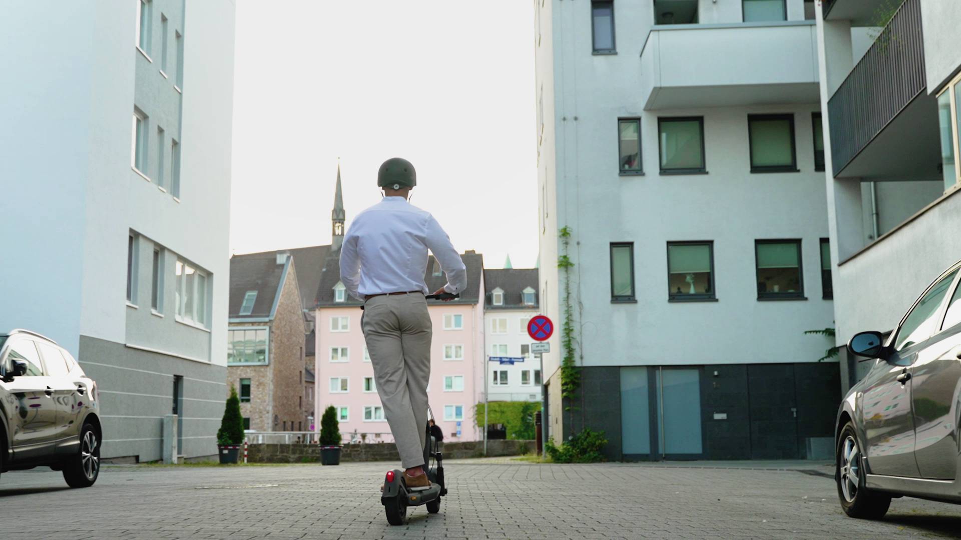 Eine Person mit Helm fährt auf einem Elektroroller durch eine moderne Stadtstraße mit hohen Wohngebäuden im Hintergrund.