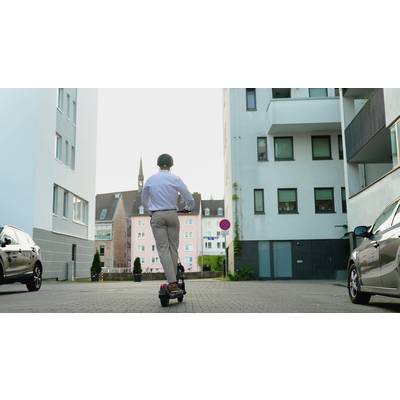 Eine Person mit Helm fährt auf einem Elektroroller durch eine moderne Stadtstraße mit hohen Wohngebäuden im Hintergrund.