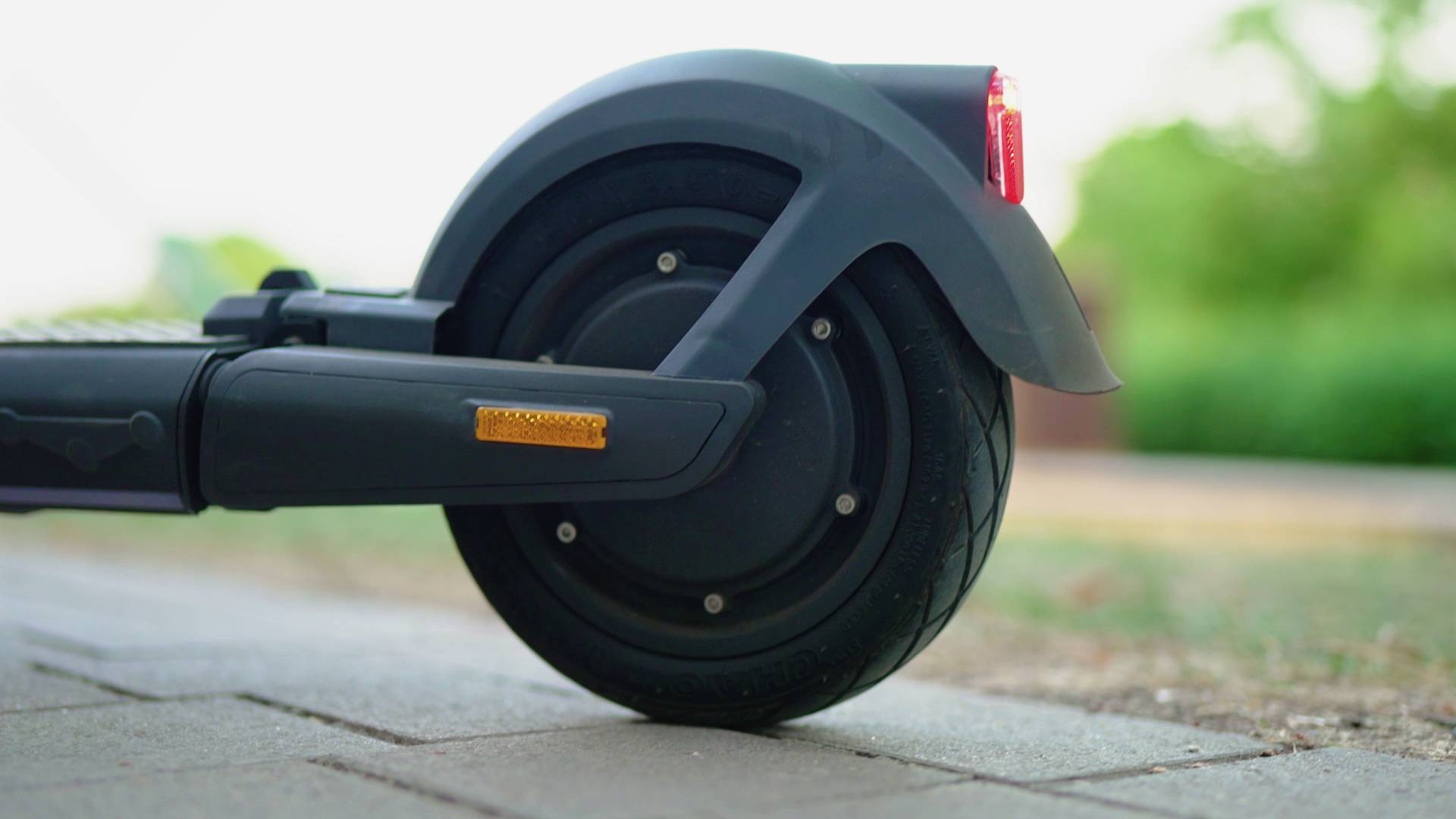 Ein elektrischer Scooter steht auf einem Gehweg. Der Fokus liegt auf dem Hinterrad mit Bremslicht und Straßenumgebung im Hintergrund.