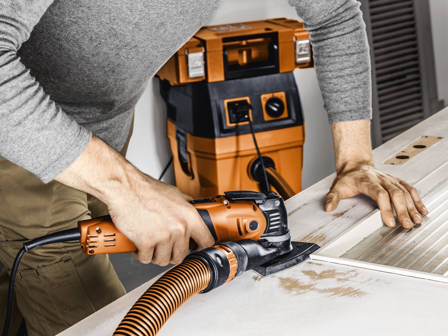 Ein Handwerker schleift eine Holzoberfläche mit einem orangen Schleifgerät. Im Hintergrund steht ein orangefarbener Staubsauger.