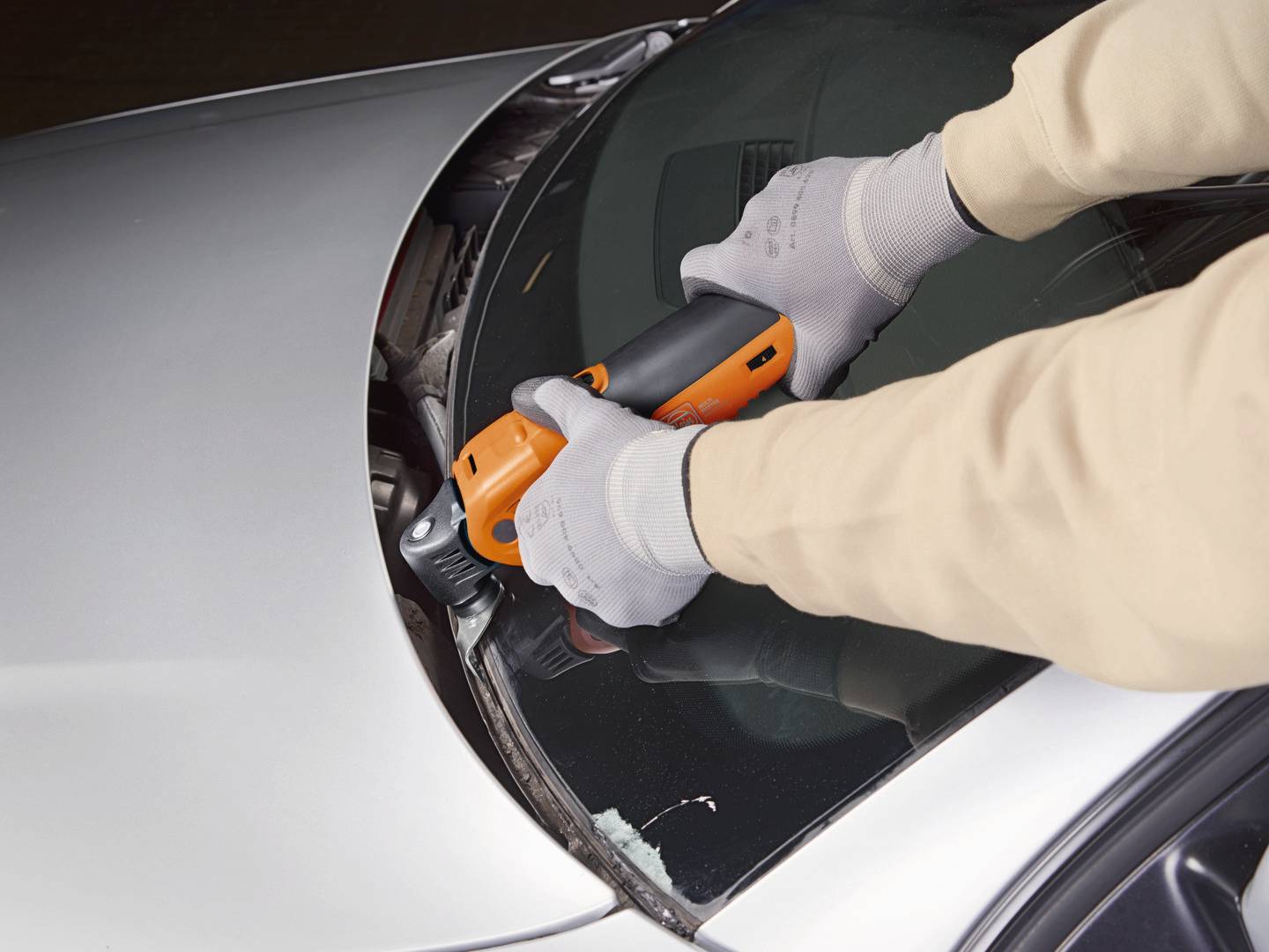 Eine Person trägt graue Handschuhe und verwendet ein orange-schwarzes Elektrowerkzeug, um die Windschutzscheibe eines Autos zu entfernen.