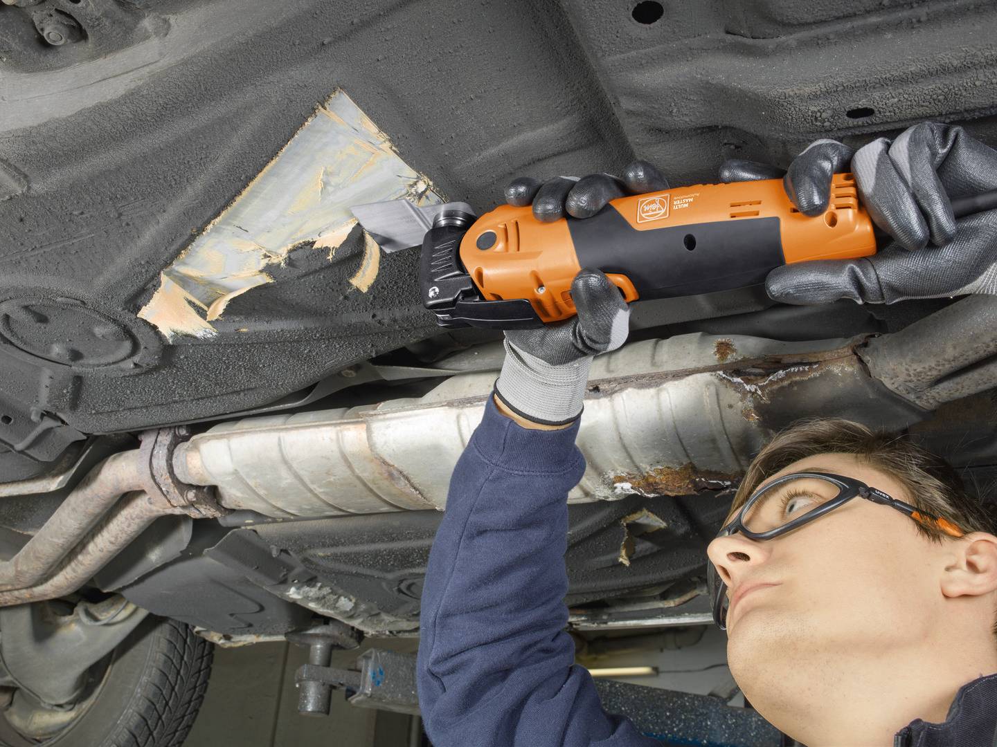 Eine Person repariert den Unterboden eines Fahrzeugs mit einem elektrischen Werkzeug. Schutzbrille und Handschuhe werden getragen.
