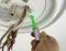 Hand hält Spannungsprüfer an Elektroanschluss in Deckenleuchte. Mehrere Kabel sind sichtbar. Gerät überprüft Stromfluss.