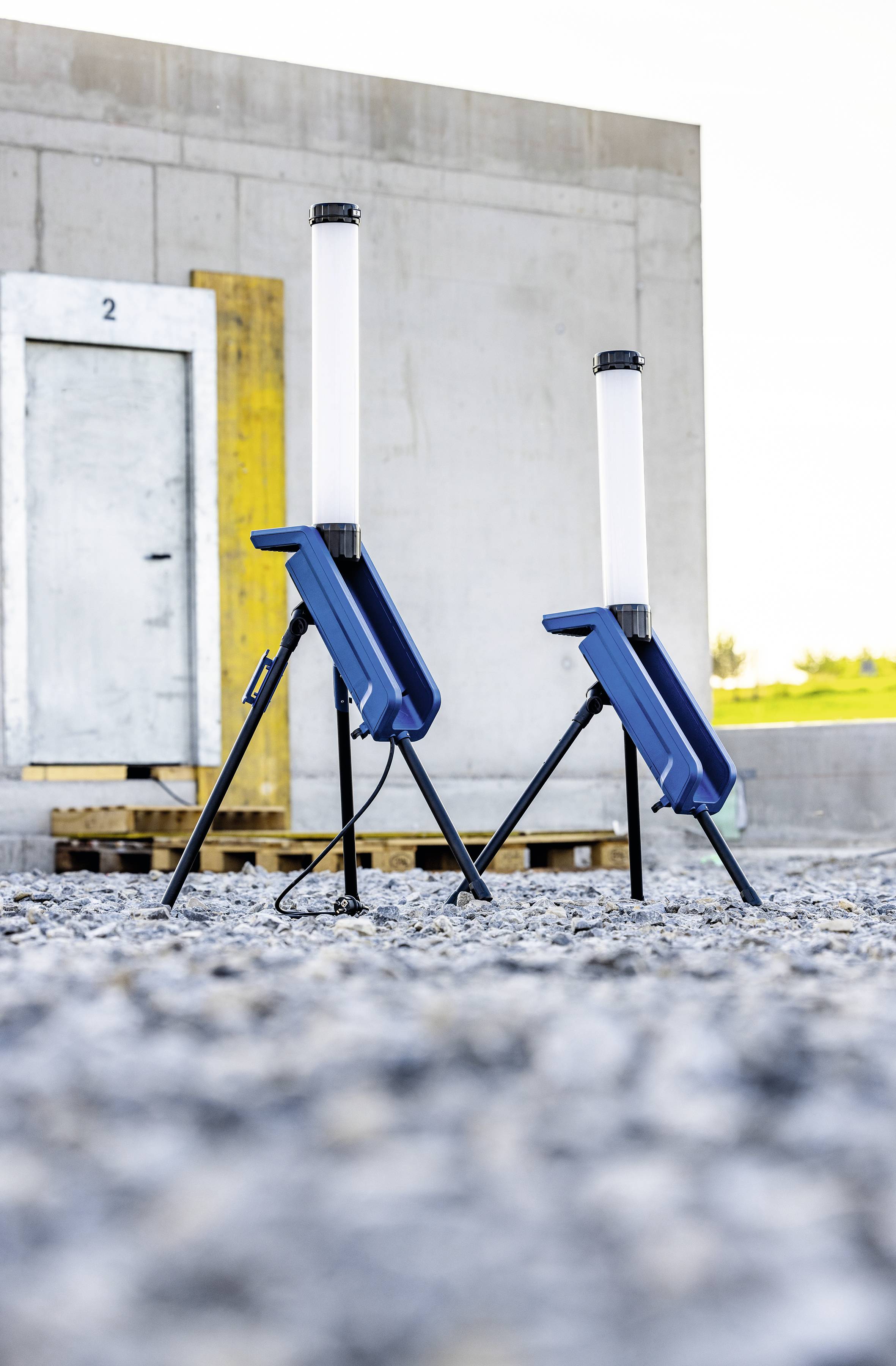 Zwei tragbare Baustrahler stehen auf einem Kiesboden vor einem Industriegebäude mit einer weißen Tür.