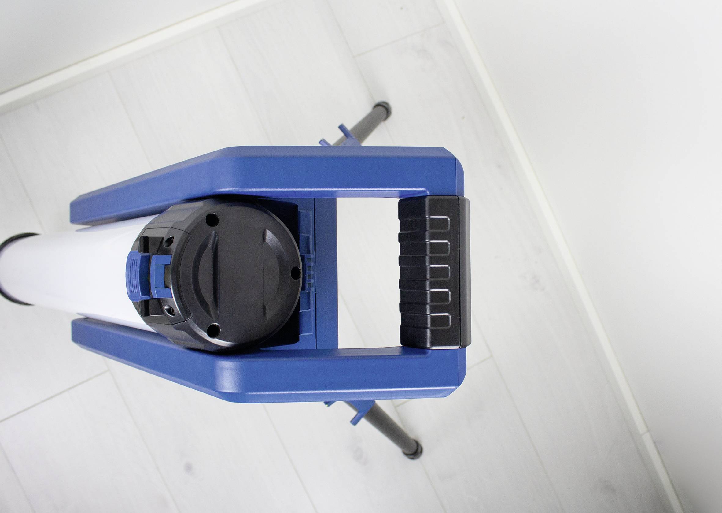 Das Bild zeigt die Oberseite eines Stativs mit einem blauen Griff und einer zylindrischen Halterung auf einem hellen Boden.