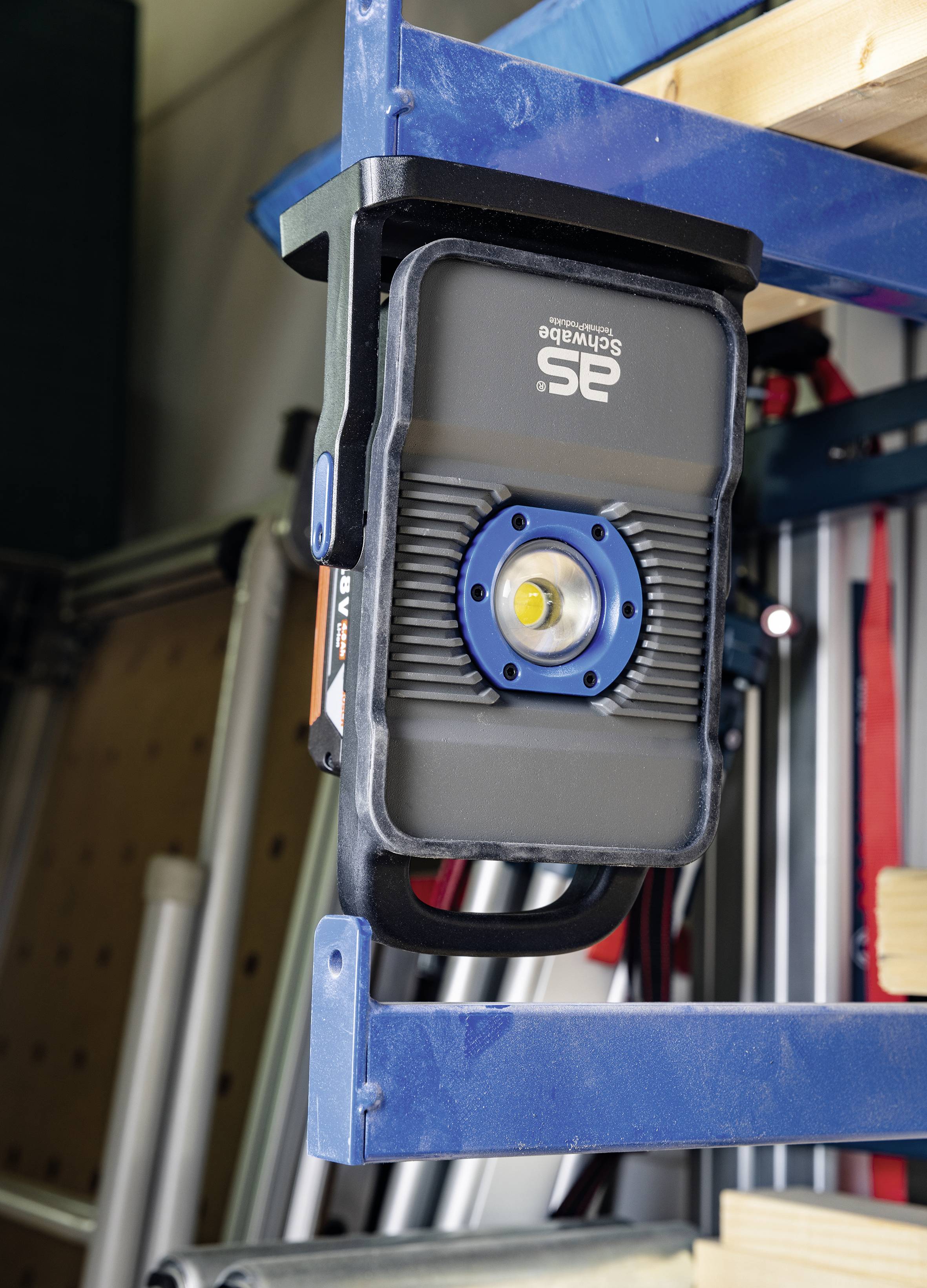'12V Akku-LED-Baustrahler' auf Werkbank in Werkstatt platziert. Im Hintergrund sind Regale mit Werkzeugen sichtbar.