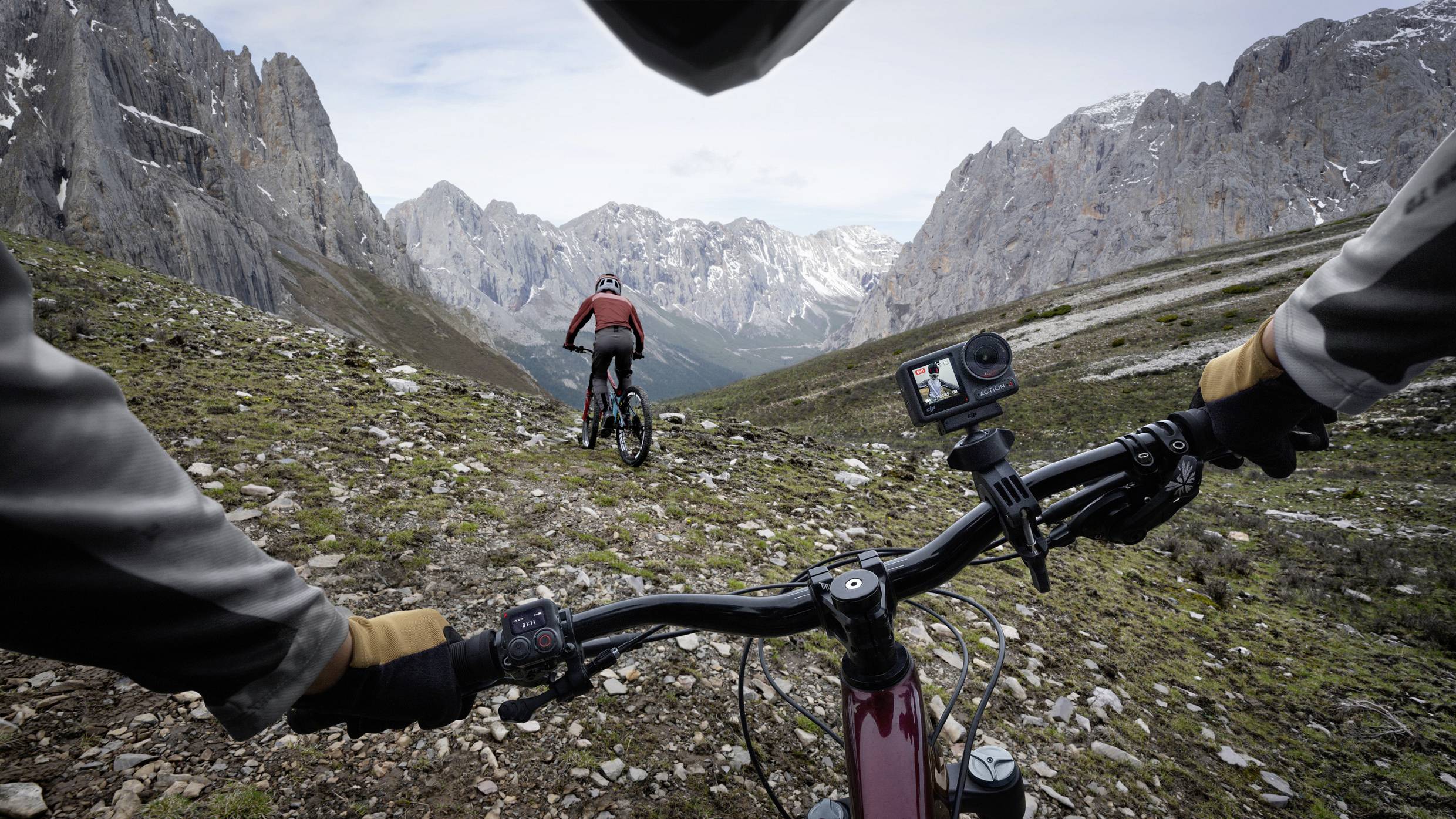 Eine Person fährt mit dem Mountainbike auf einem steinigen Pfad in bergiger Landschaft, gefolgt von einer anderen in der Ferne.