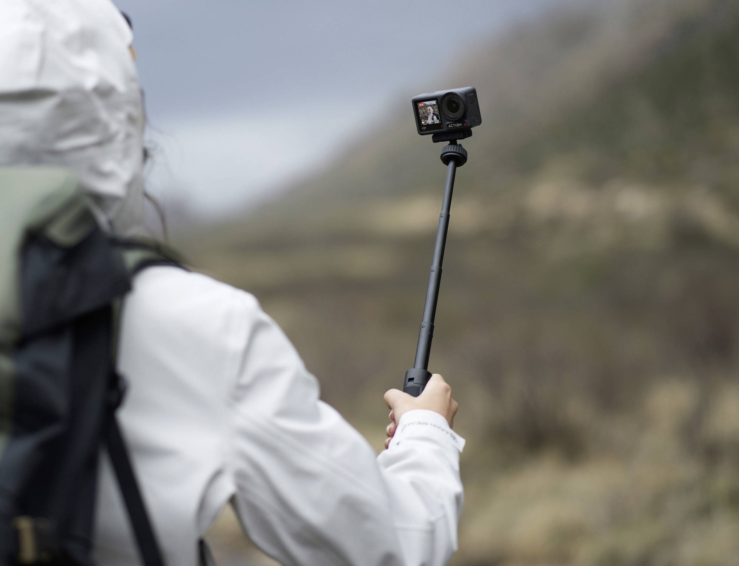 Eine Person in Outdoor-Kleidung hält eine Action-Kamera an einem Selfie-Stick in einer bergigen Landschaft.