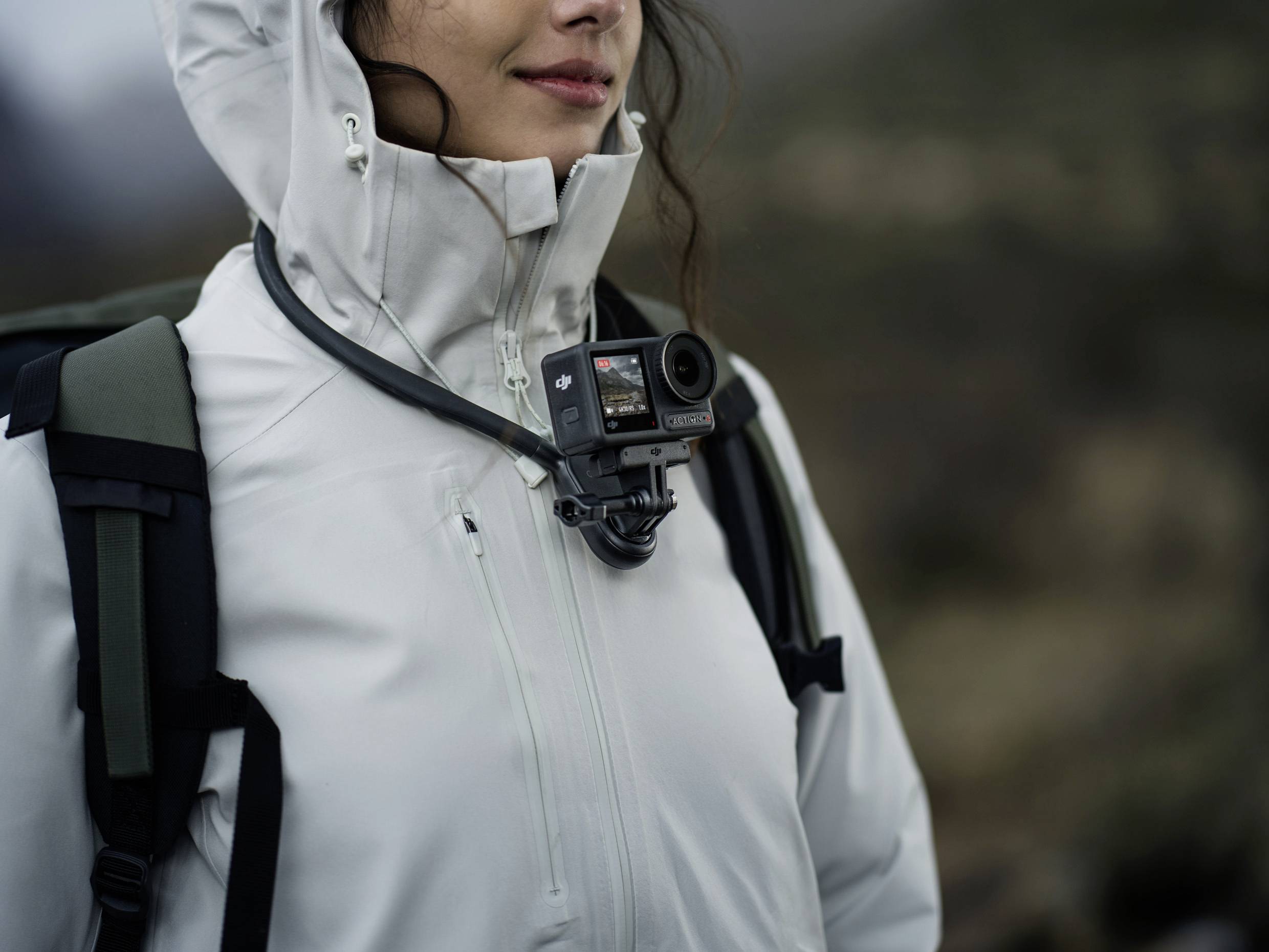 Person mit weißer Jacke und Rucksack trägt eine Action-Kamera auf Brusthöhe in einer bergigen Landschaft.