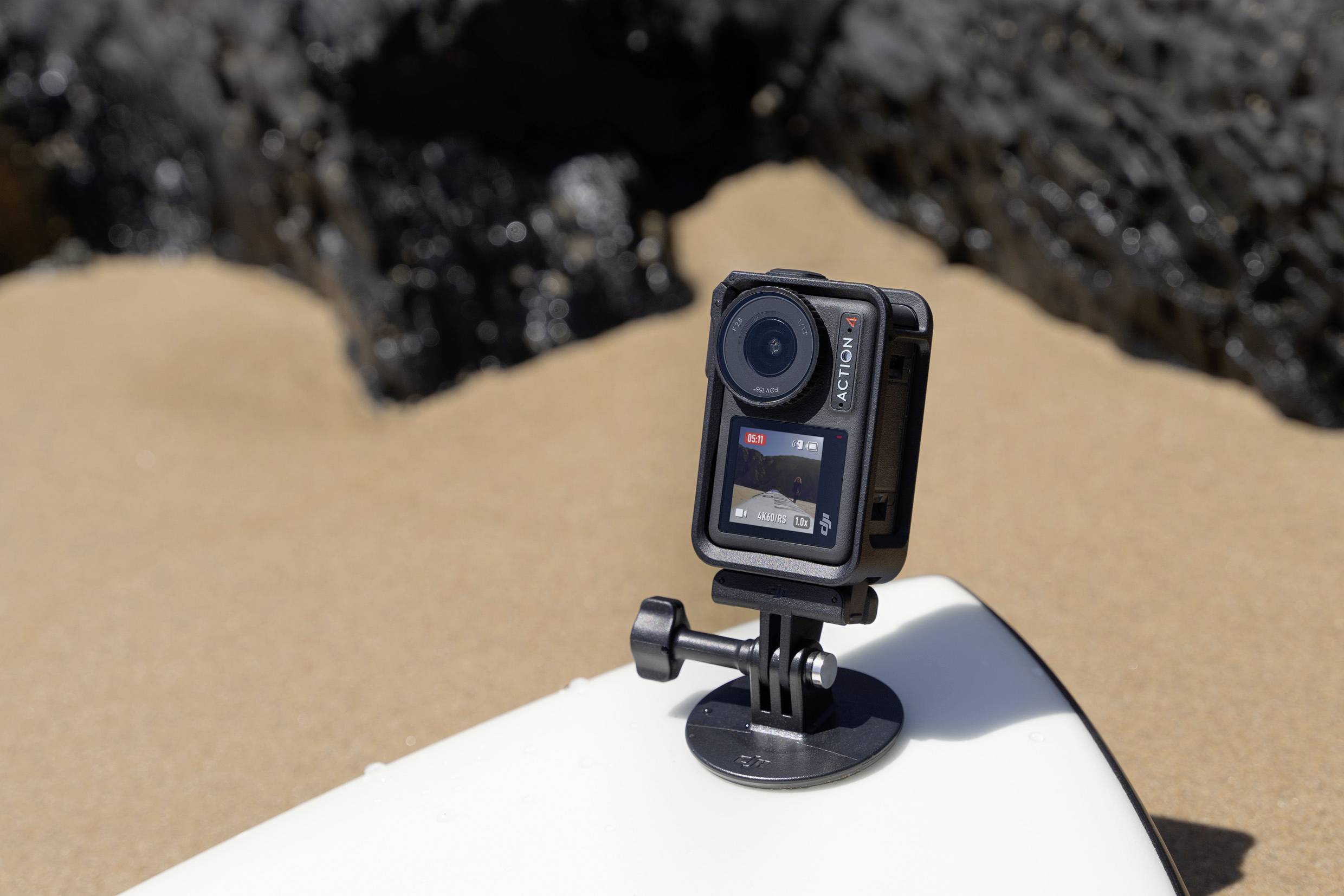 Eine Action-Kamera ist auf einem Surfbrett montiert, das auf sandigem Boden liegt. Im Hintergrund sind dunkle Felsen sichtbar.