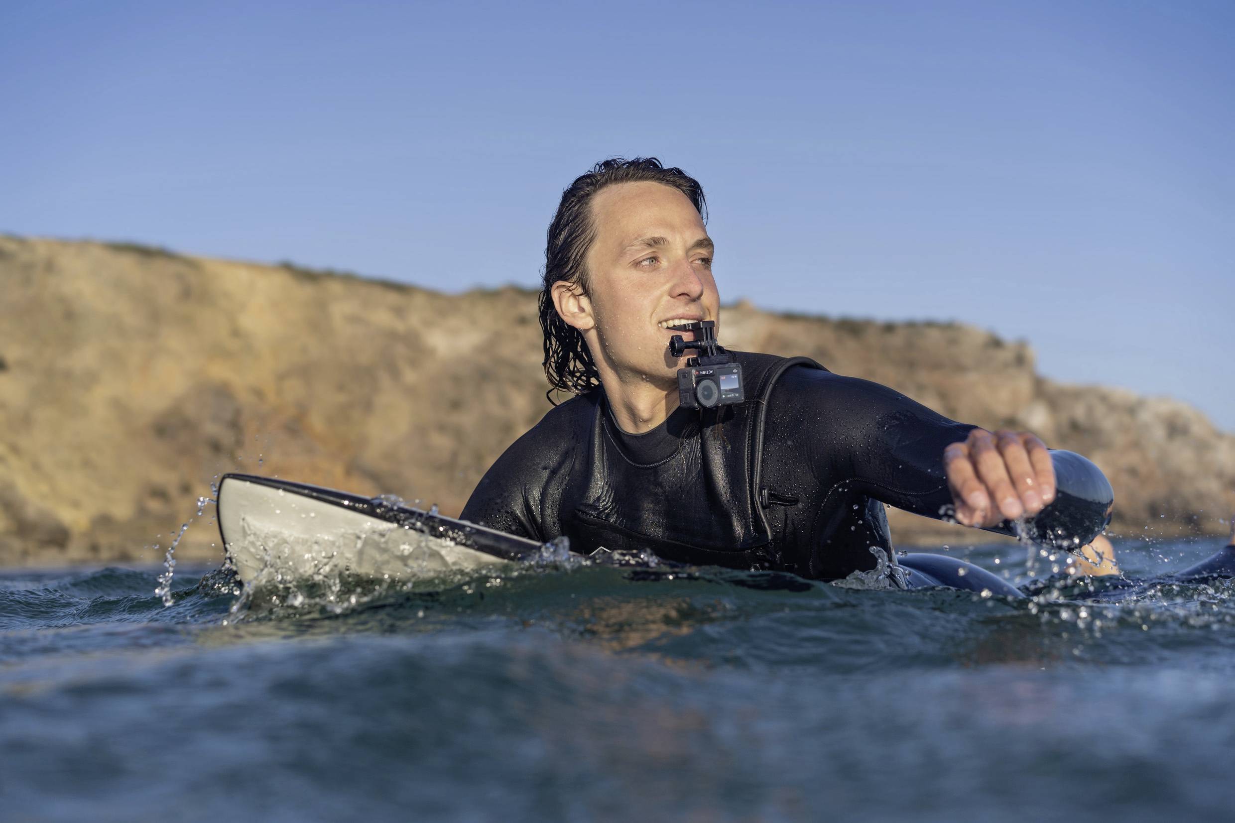 Eine Person in einem Neoprenanzug paddelt auf einem Surfbrett im Meer. Im Hintergrund ist eine felsige Küste zu sehen.