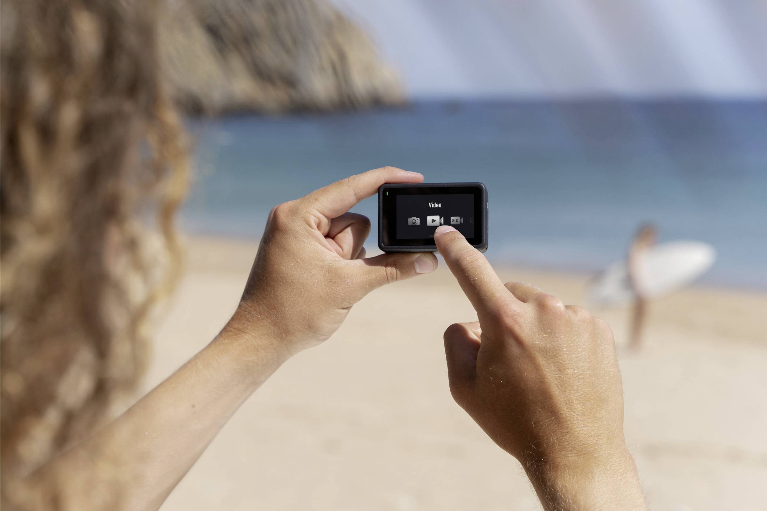Eine Person hält eine Action-Kamera am Strand. Im Hintergrund steht jemand mit einem Surfbrett am Wasser.