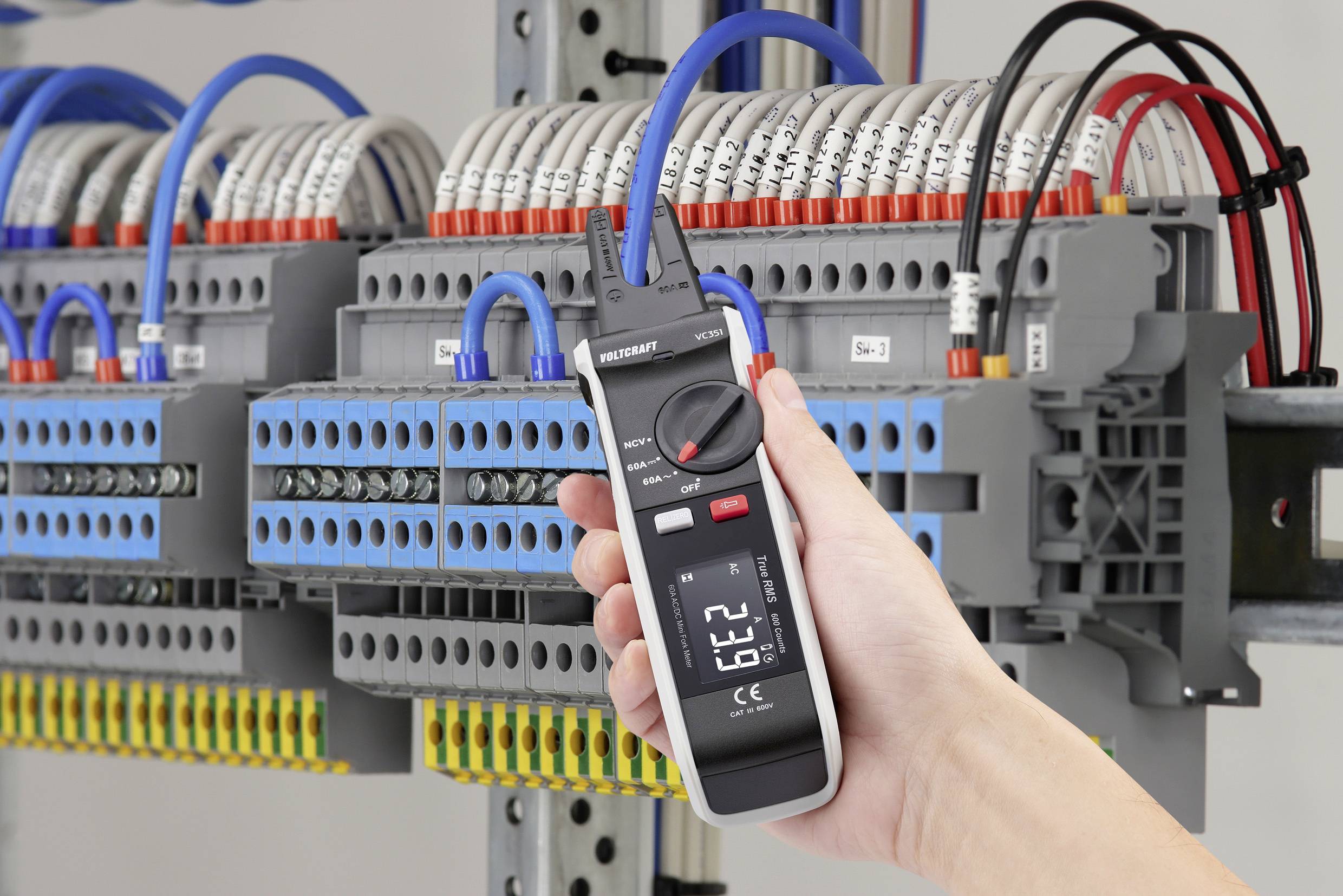 Eine Hand hält ein digitales Multimeter, das den Stromfluss in einem Schaltschrank misst. Im Hintergrund sind Kabel und Anschlüsse sichtbar.