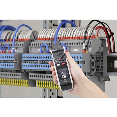 Eine Hand hält ein digitales Multimeter, das den Stromfluss in einem Schaltschrank misst. Im Hintergrund sind Kabel und Anschlüsse sichtbar.