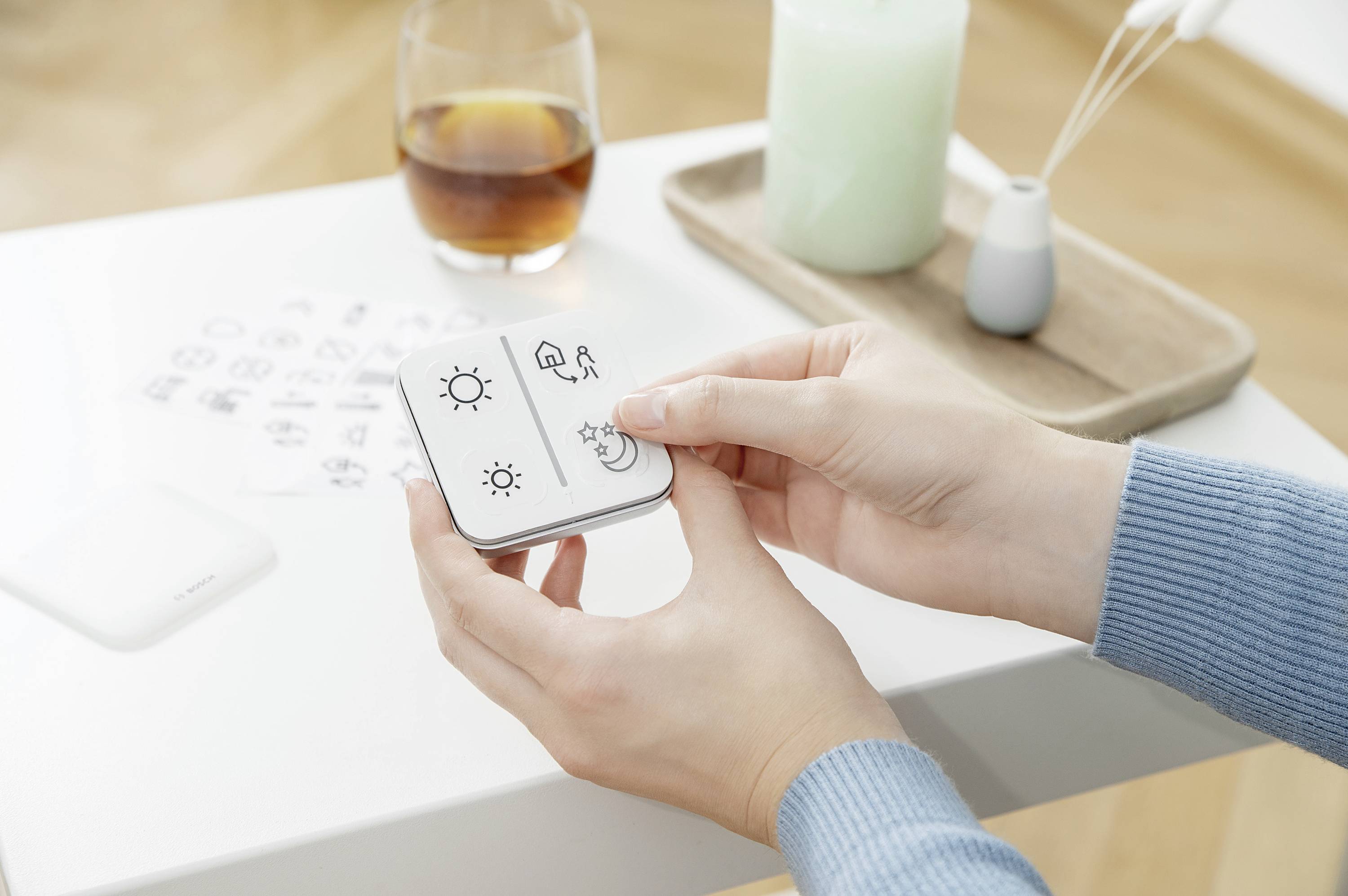 Eine Person hält ein kleines Gerät mit Symbolen, darunter eine Sonne und ein Mond. Daneben stehen ein Getränk und eine Kerze auf einem Tisch.